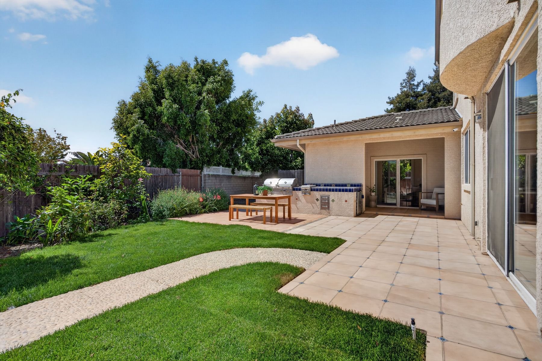 Backyard patio with tiled flooring, green lawn, garden area with bushes, a tree, wooden table with benches, built-in grill, and a sliding glass door leading into the house under a blue sky.
