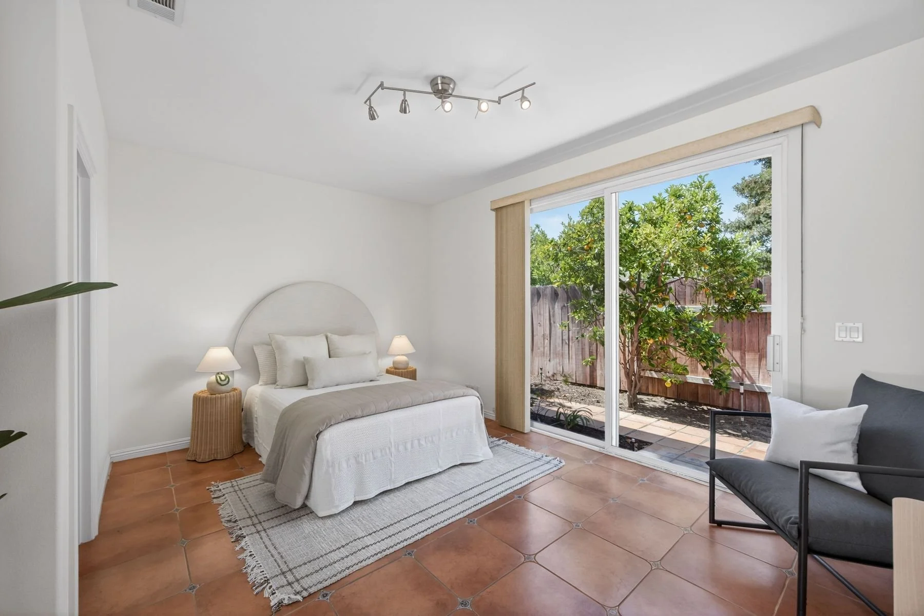 A bright bedroom with a white bed, twin nightstands with lamps, a sliding glass door opening to a backyard with a tree, and a gray sofa.