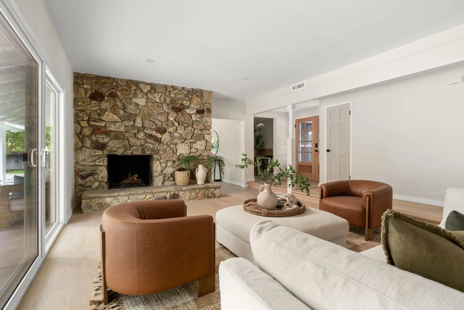 Living room with a stone fireplace, sliding glass door, tan wooden floor, white walls, a white couch, brown leather chairs, a coffee table with a vase and greenery, and a wooden door.