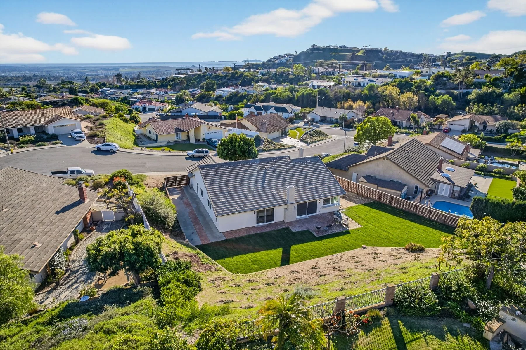 Aerial view of a suburban neighborhood with single-family homes, green lawns, trees, and a swimming pool, with hills and a distant coastline in the background.
