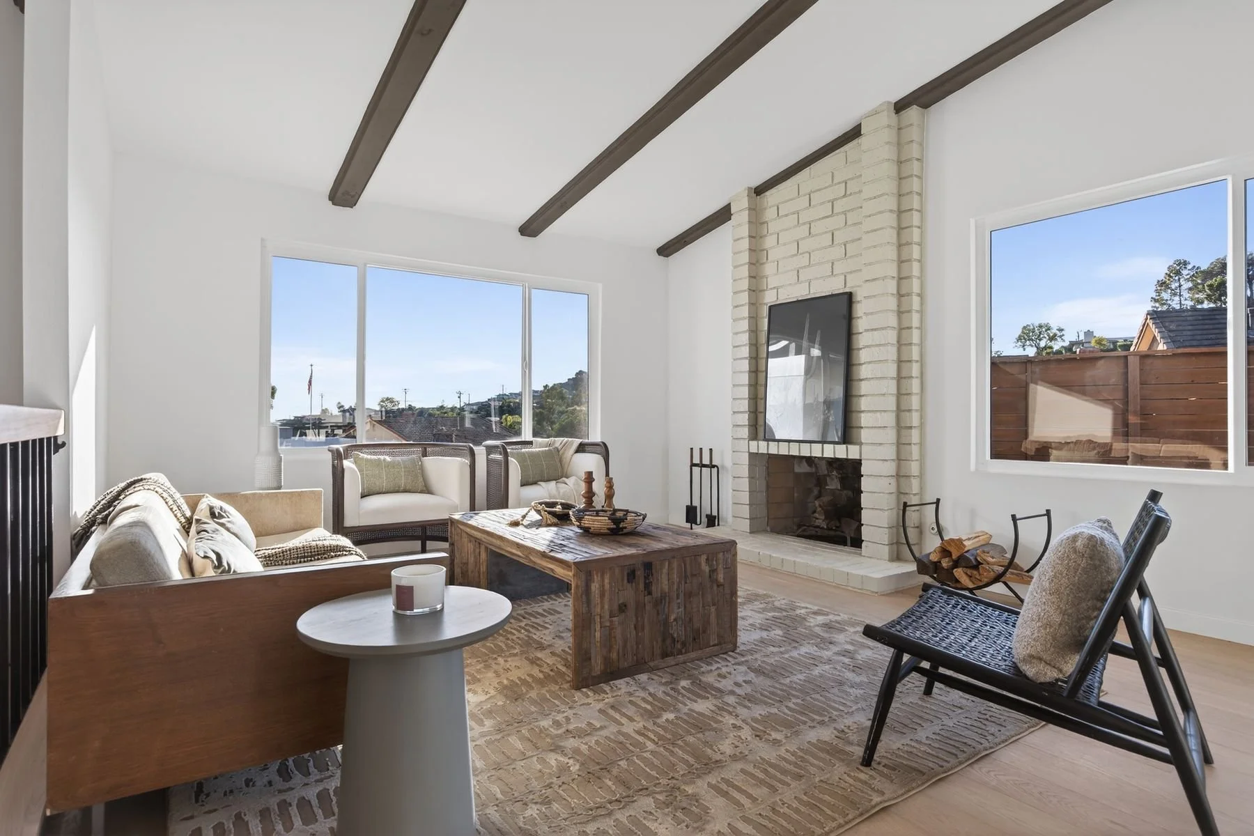 Bright living room with white brick fireplace, large windows, wooden beams on ceiling, and mix of modern and rustic furniture.