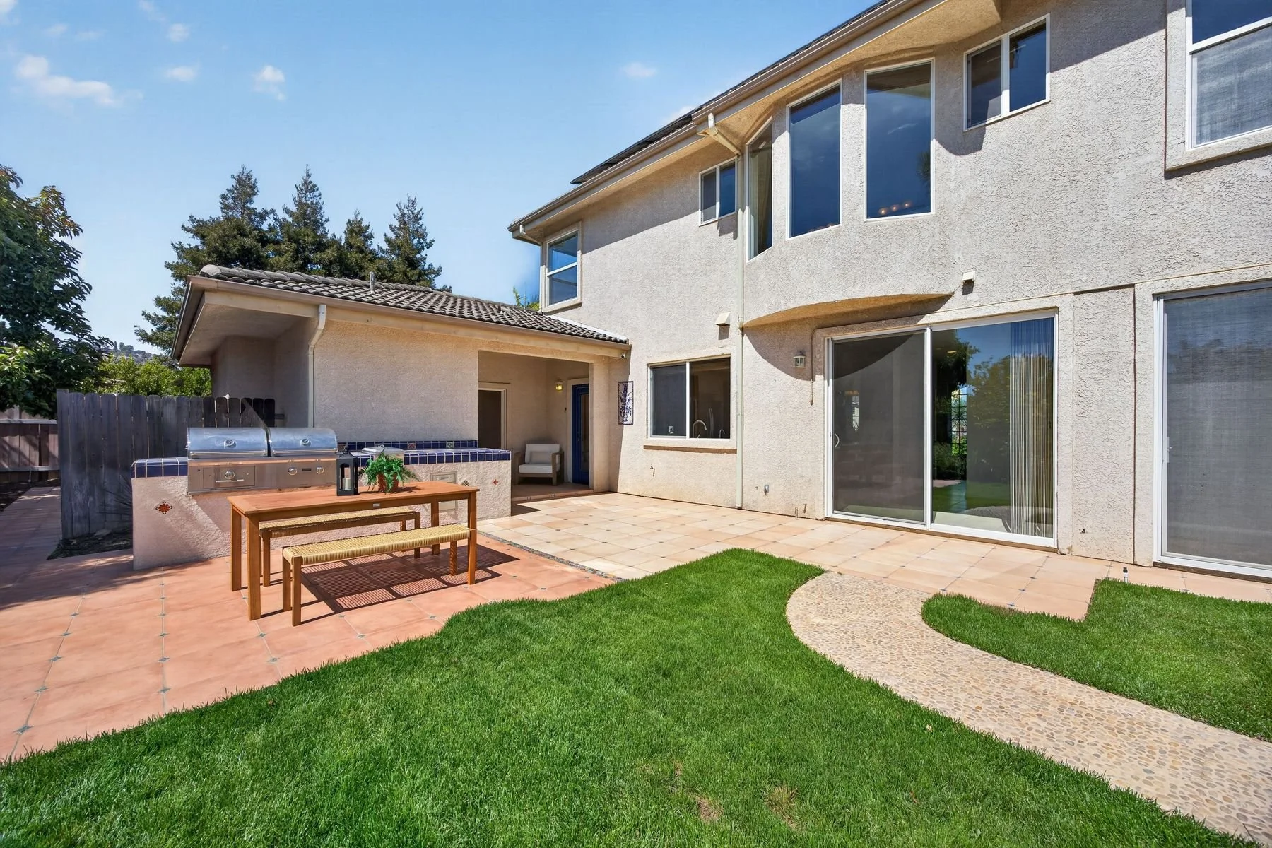 Backyard area of a house with a green lawn, tiled patio, outdoor kitchen with grilling station, a sliding glass door, and large windows on the house wall under a clear blue sky.