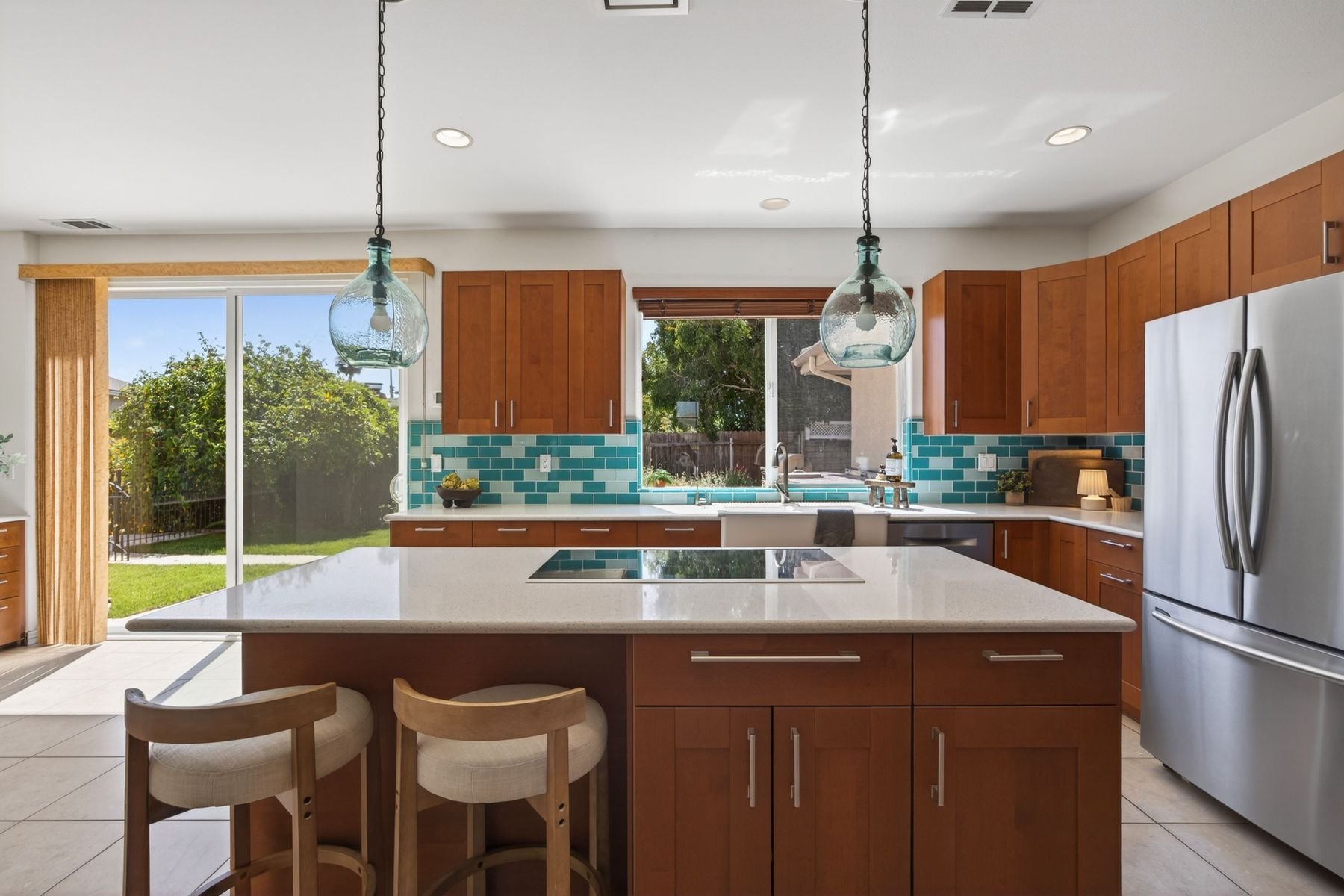 Modern kitchen with wooden cabinets, a white island, two glass pendant lights, a stainless steel refrigerator, and large windows showing a backyard with green grass and trees.