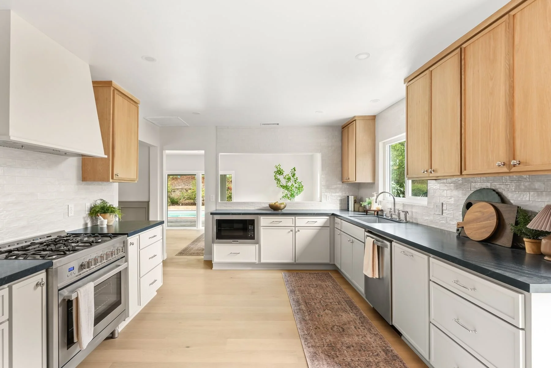 Bright modern kitchen with white cabinets, black countertops, and light wood upper cabinets. Features a gas stove, microwave, double sink, and decorative plants. Large window and sliding door reveal a backyard with greenery and pool.