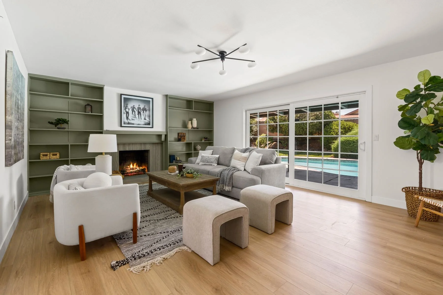 Living room with a fireplace, gray sofa, white armchairs, sliding glass door, green bookshelves, potted tree, and wooden coffee table.