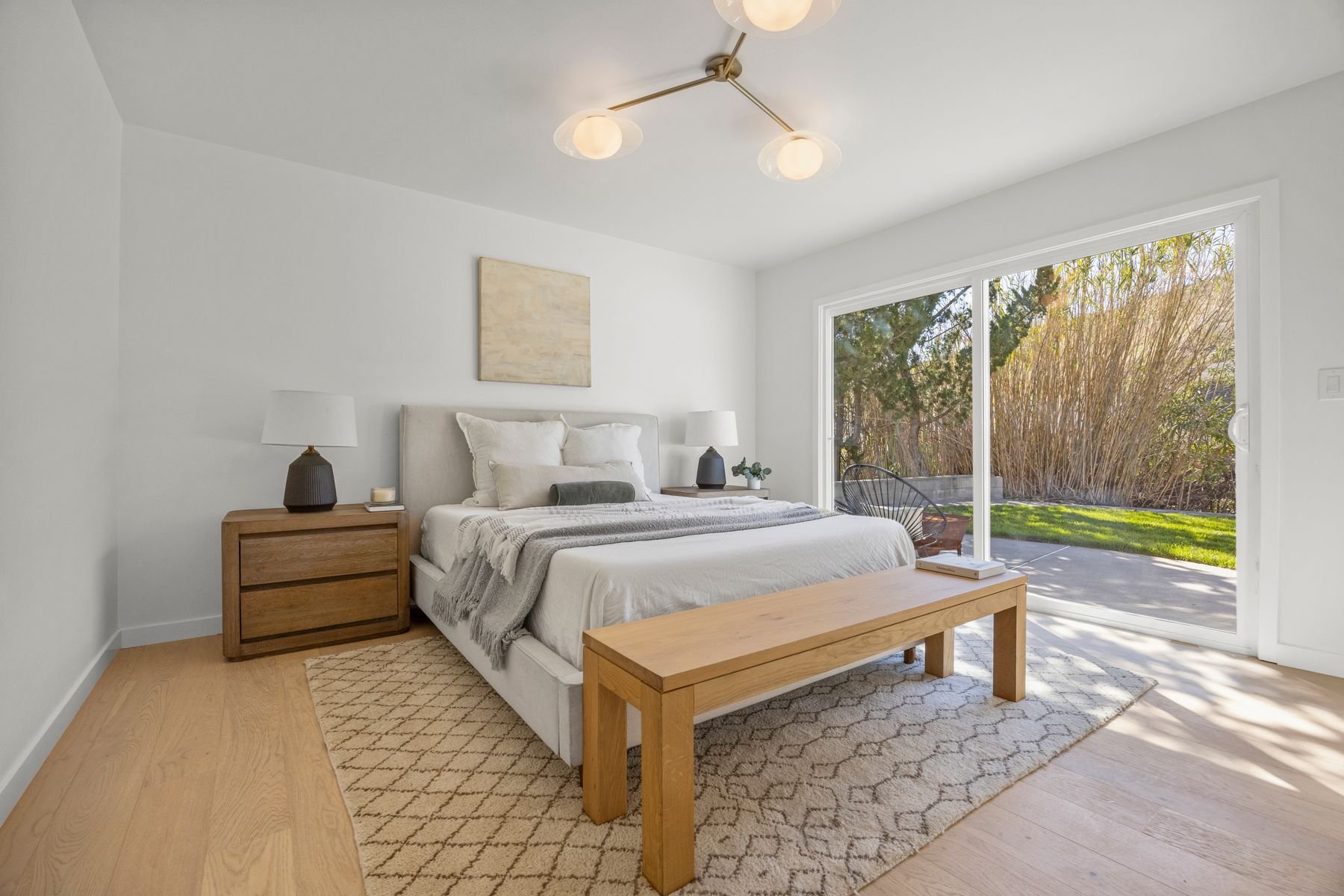 Bright and contemporary bedroom with white walls, a large sliding glass door opening to an outdoor scene with trees and grass, a queen-sized bed with white linens and plush pillows, two wooden nightstands with lamps, a beige rug, a wooden bench at th