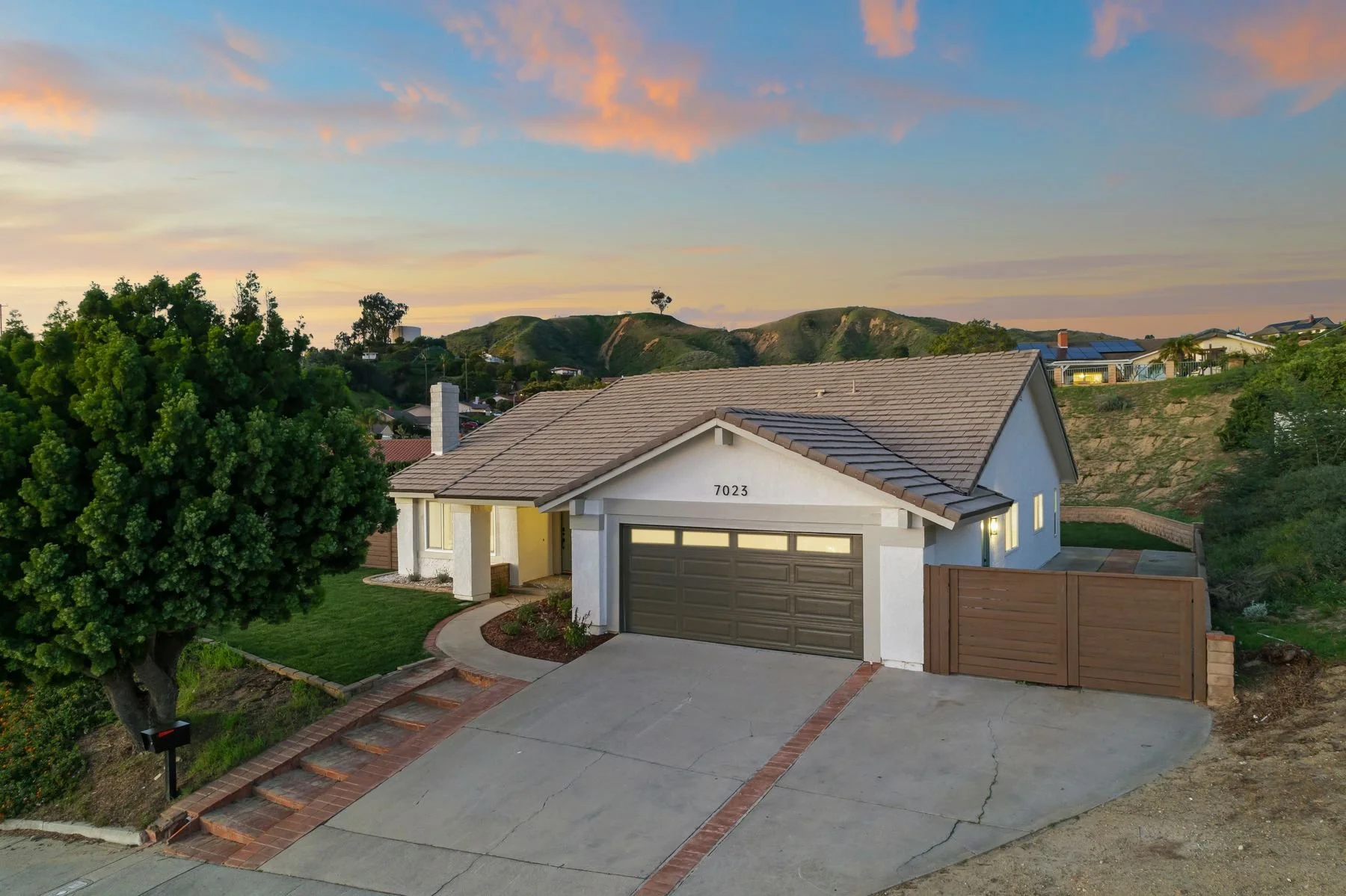 A modern single-story house with a gray garage door, white walls, and a tiled roof. The house number '7023' is visible. There is a large green tree to the left of the driveway, and a curved walkway bordered by bricks leads to the front entrance. The 