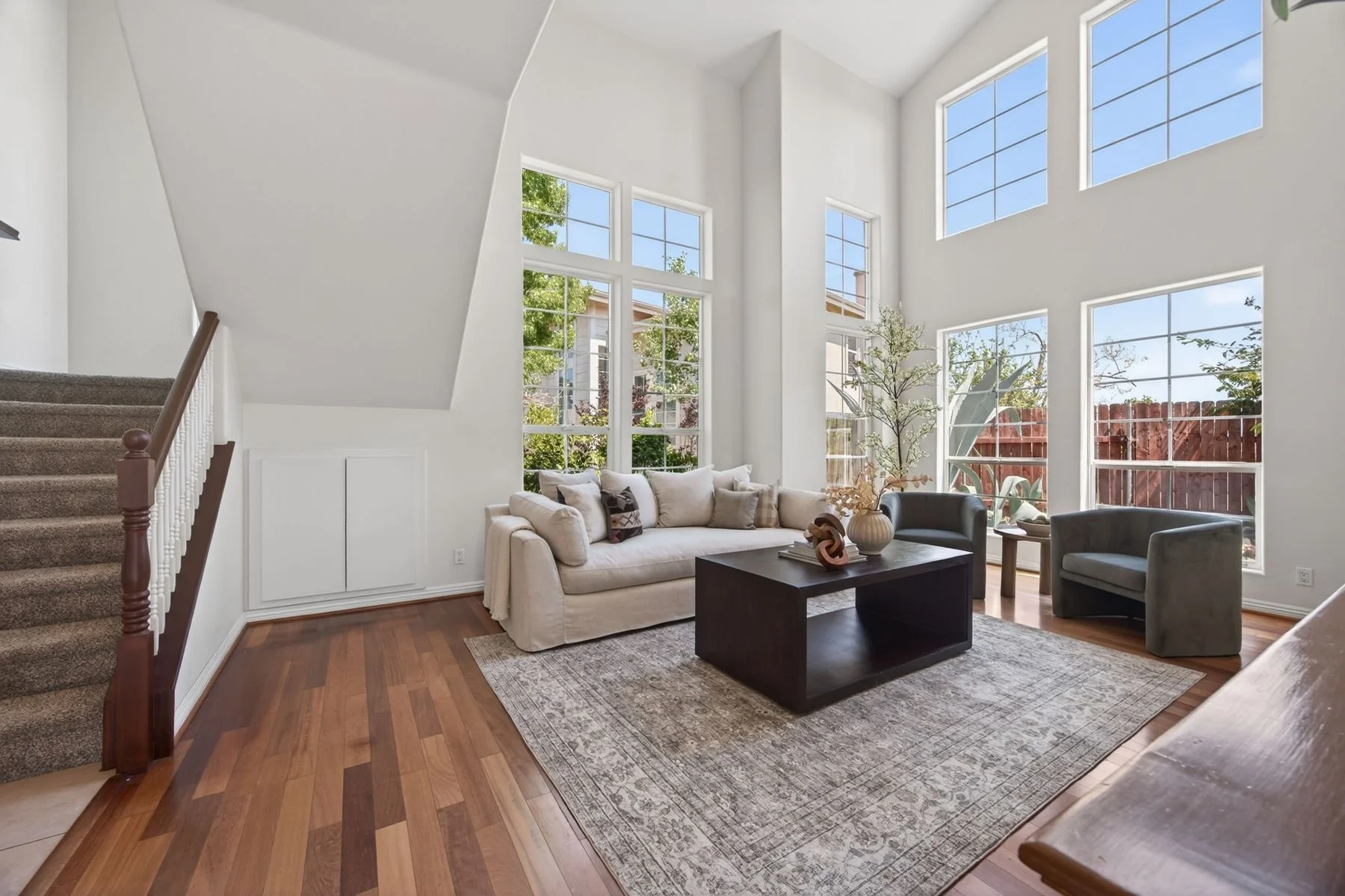 Bright living room with large windows, white walls, and hardwood floors. Contains a white sofa with multiple pillows, a dark coffee table, two armchairs, and decorative vases with plants. Carpet underneath the coffee table and staircase with carpeted