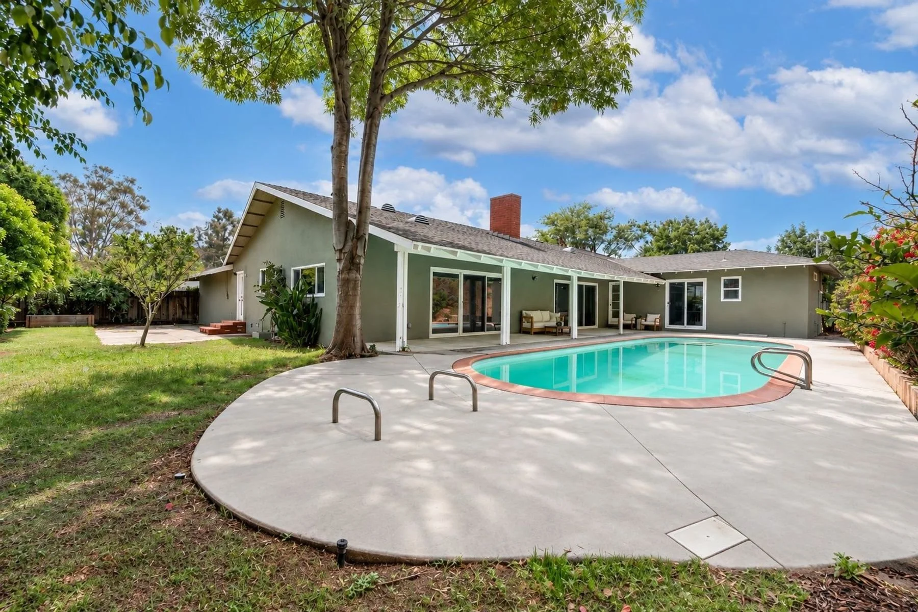 Backyard with a kidney-shaped swimming pool, surrounding concrete deck, green lawn, trees, and a single-story house with sliding glass doors and a covered patio.