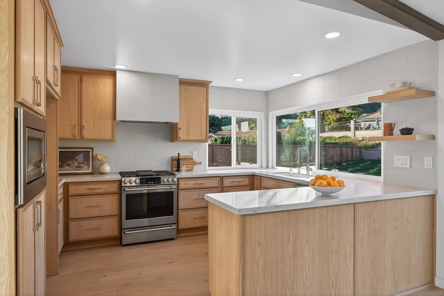 Modern kitchen with wooden cabinets, white marble countertops, stainless steel oven, and large windows overlooking a backyard with a fence and trees. A bowl of oranges is on the kitchen island, and open shelves hold bowls and a teapot.