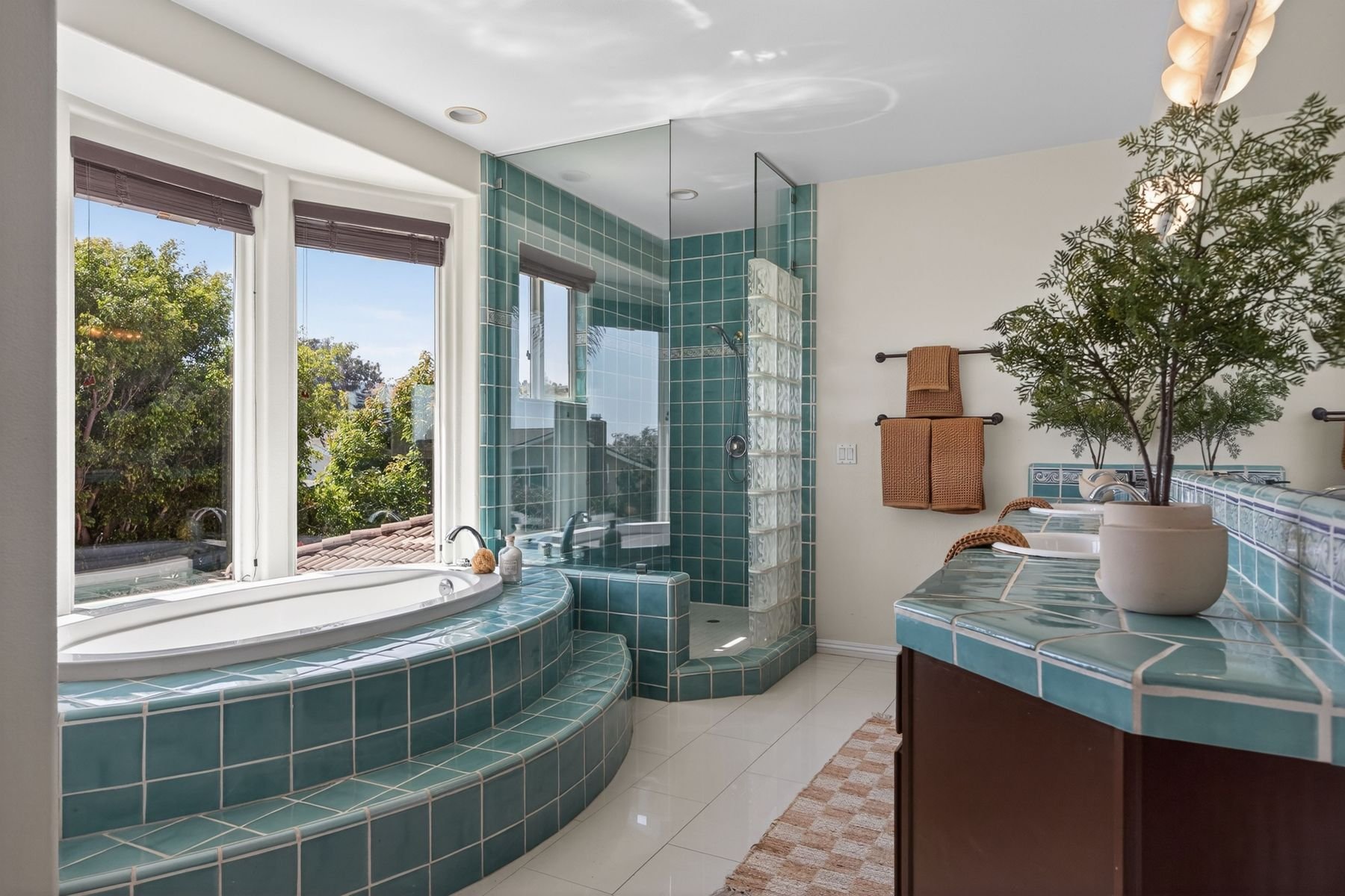 A bathroom with a large corner bathtub and a walk-in shower, both tiled in green tiles. The room has a large window with blinds and a view of trees outside. There are towels hanging on a rack and a potted plant on the tiled countertop, with a soap di