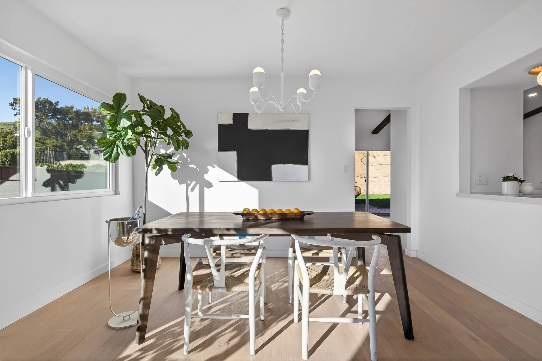 Bright dining room with white walls, large window showing outdoor trees, modern chandelier, abstract black and white artwork, wooden dining table, white chairs, a potted plant, and a track light.