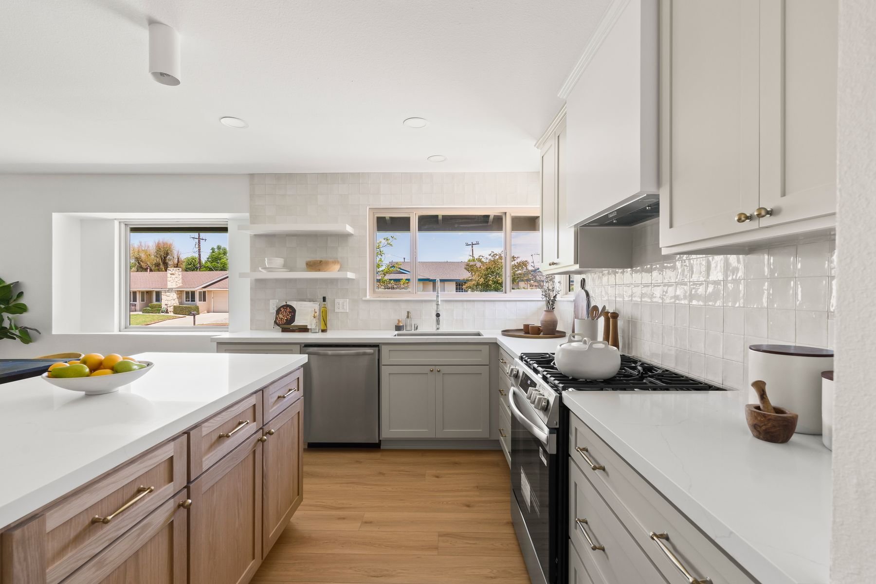 Bright, modern kitchen with white countertops and cabinets, a stainless steel stove, and a bowl of fruit on the island. Large windows show a suburban neighborhood outside.