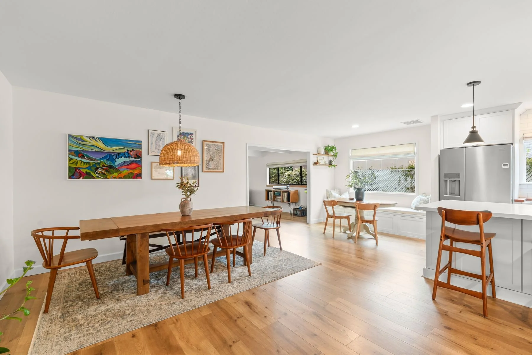 Open-concept dining and kitchen area with wooden dining table, chairs, and bar stools, artwork on white walls, hardwood floors, and large windows