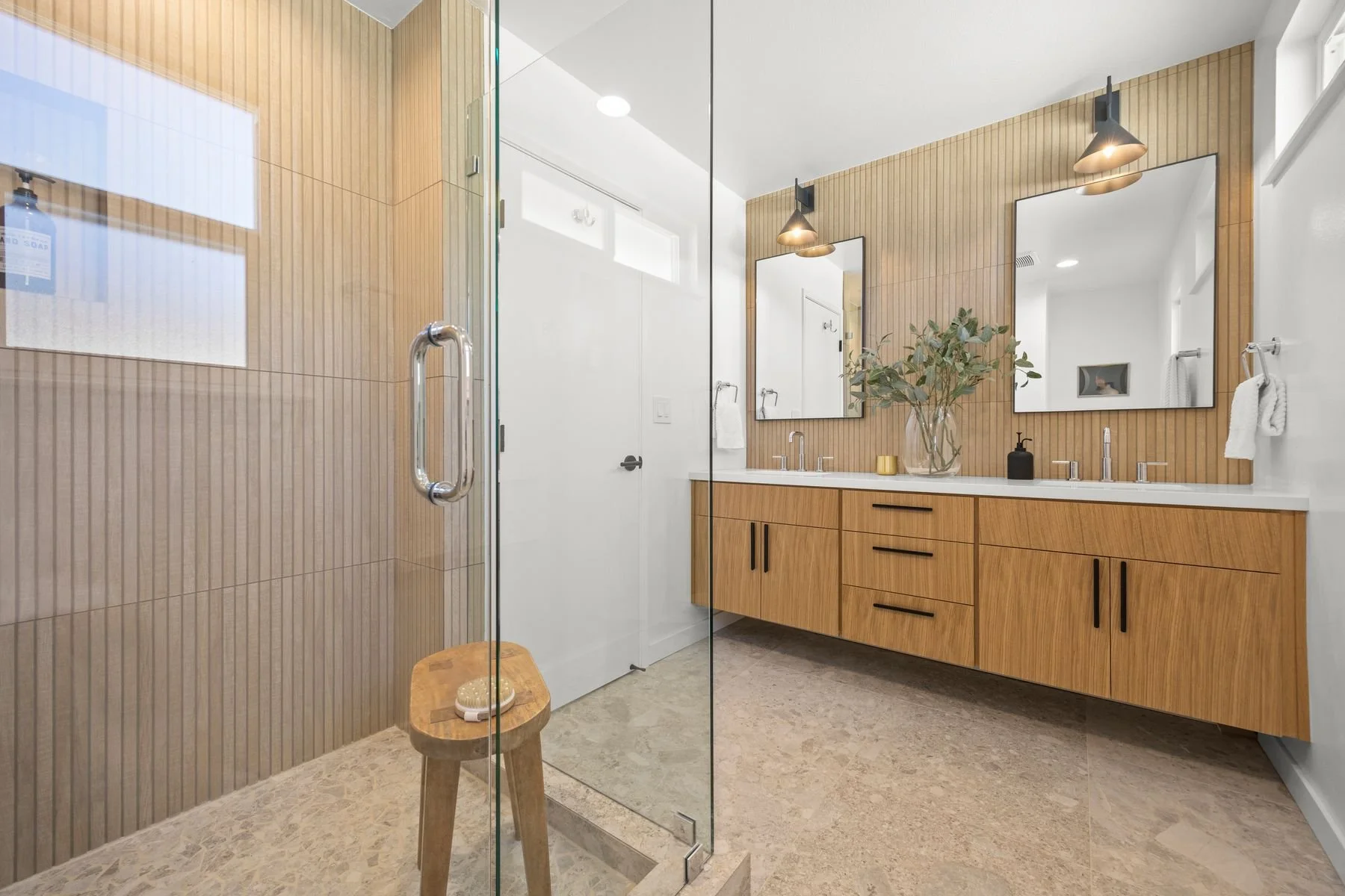 Modern bathroom with wood accents, a glass shower enclosure, a double mirror above a wooden vanity, and decorative elements like a plant, black soap dispenser, and towel.