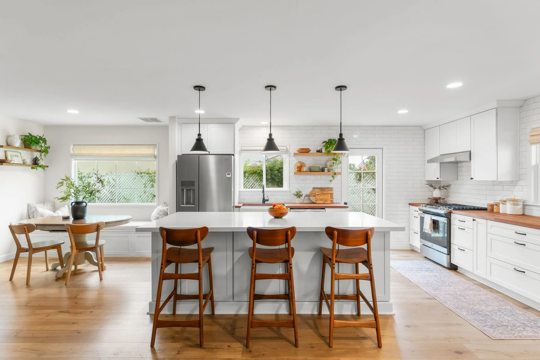 Modern kitchen with white cabinets, wooden countertops, stainless steel appliances, and a large island with a white surface and wooden chairs, light wood flooring, and a sunny window.