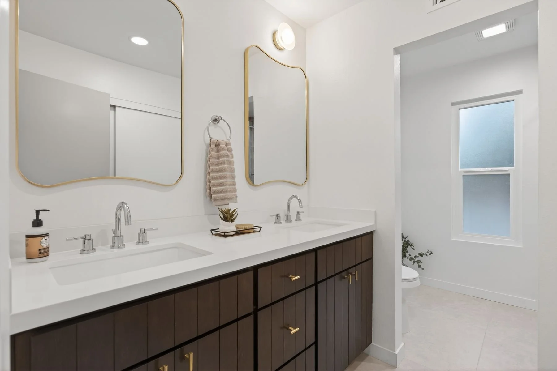 Modern bathroom with dual sinks, white countertops, dark wood cabinets, gold accents, large mirrors, a small decorative plant, and a window with natural light.