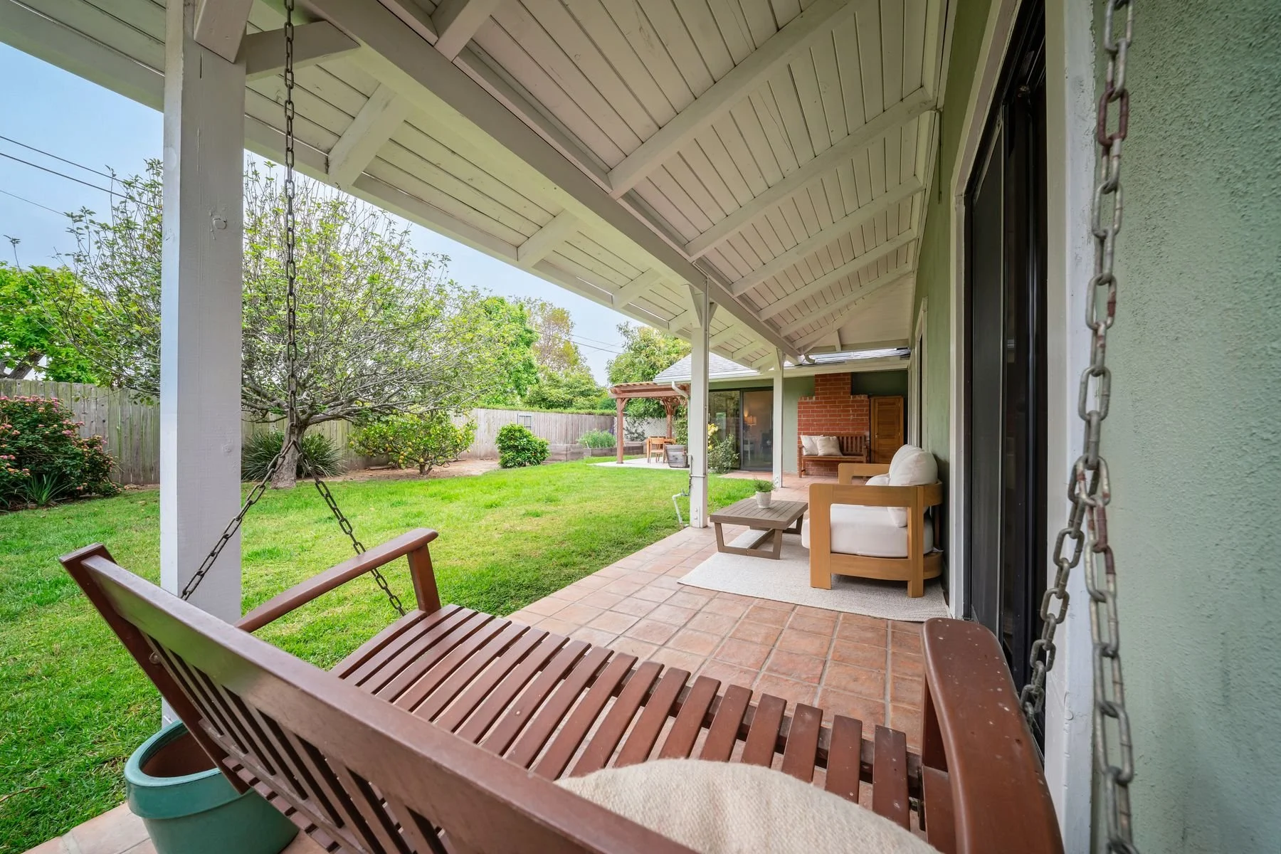 Covered backyard patio with wooden swing, cushioned armchair, small coffee table, brick floor, green lawn, trees, and garden in the background.