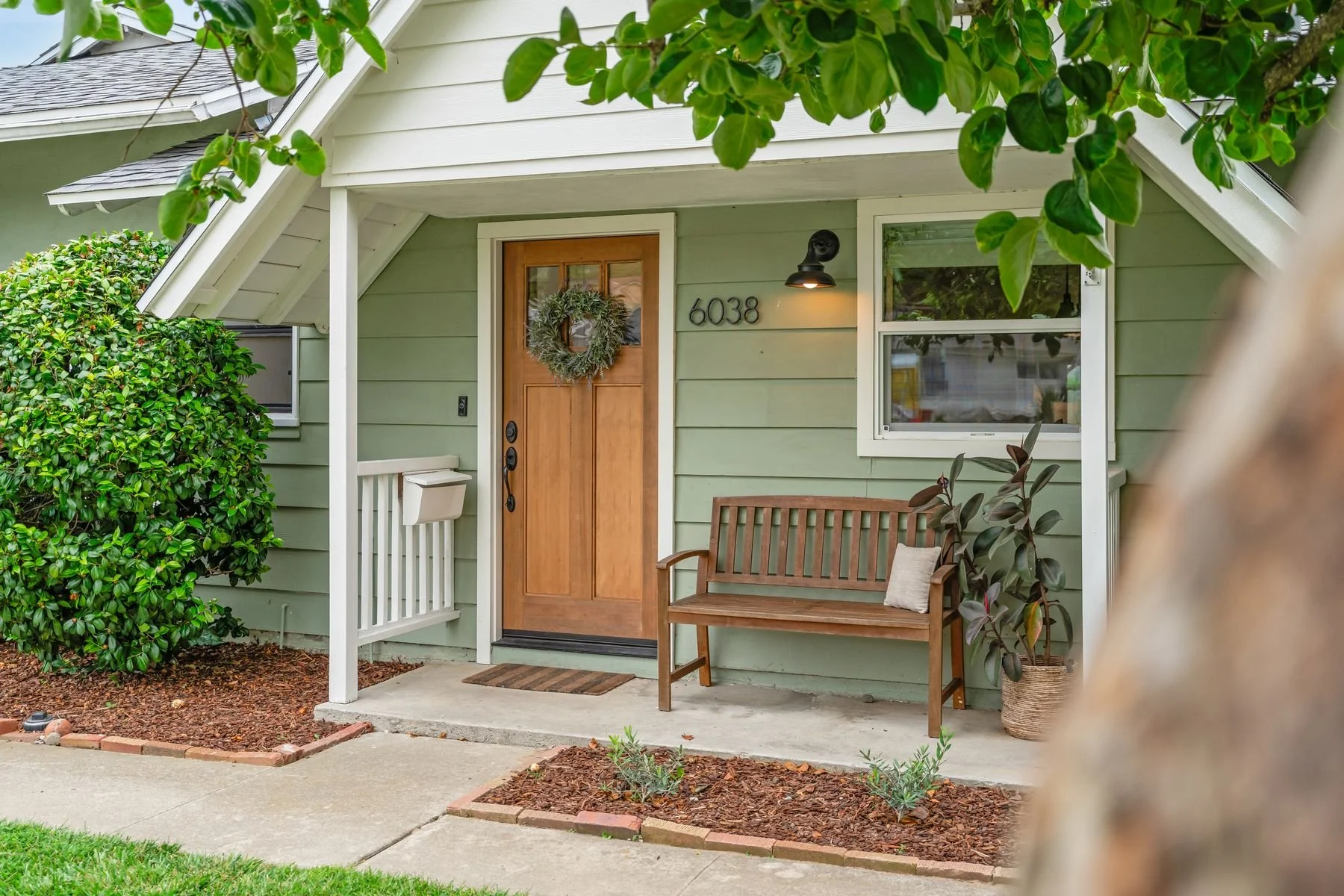 Front porch of a house with a wooden door decorated with a wreath, a black wall-mounted light, a window, a porch bench with a pillow, surrounding greenery, and landscaped garden bed.