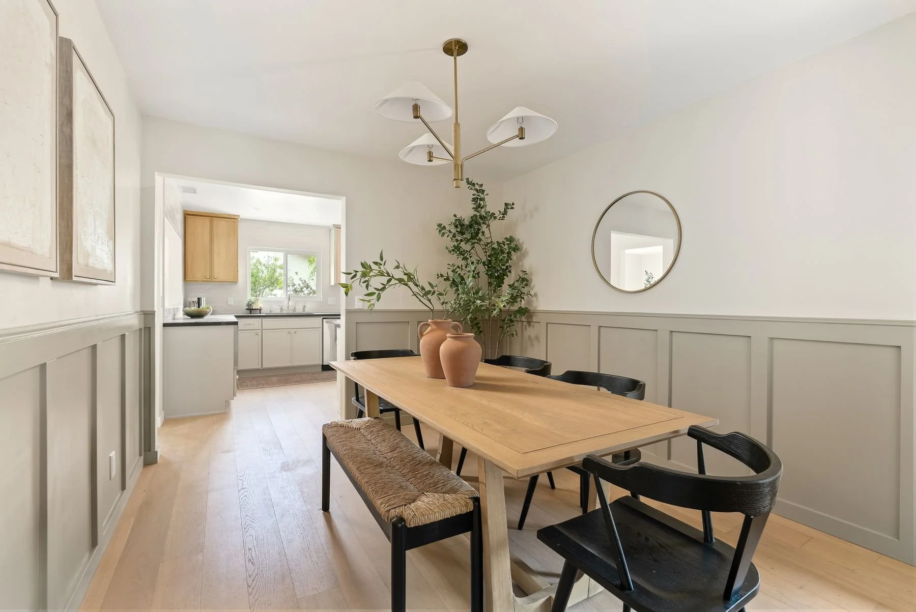 Dining room and kitchen with wooden table, black chairs, beige wainscoting, large green plant, vases, round mirror, and contemporary pendant lights.
