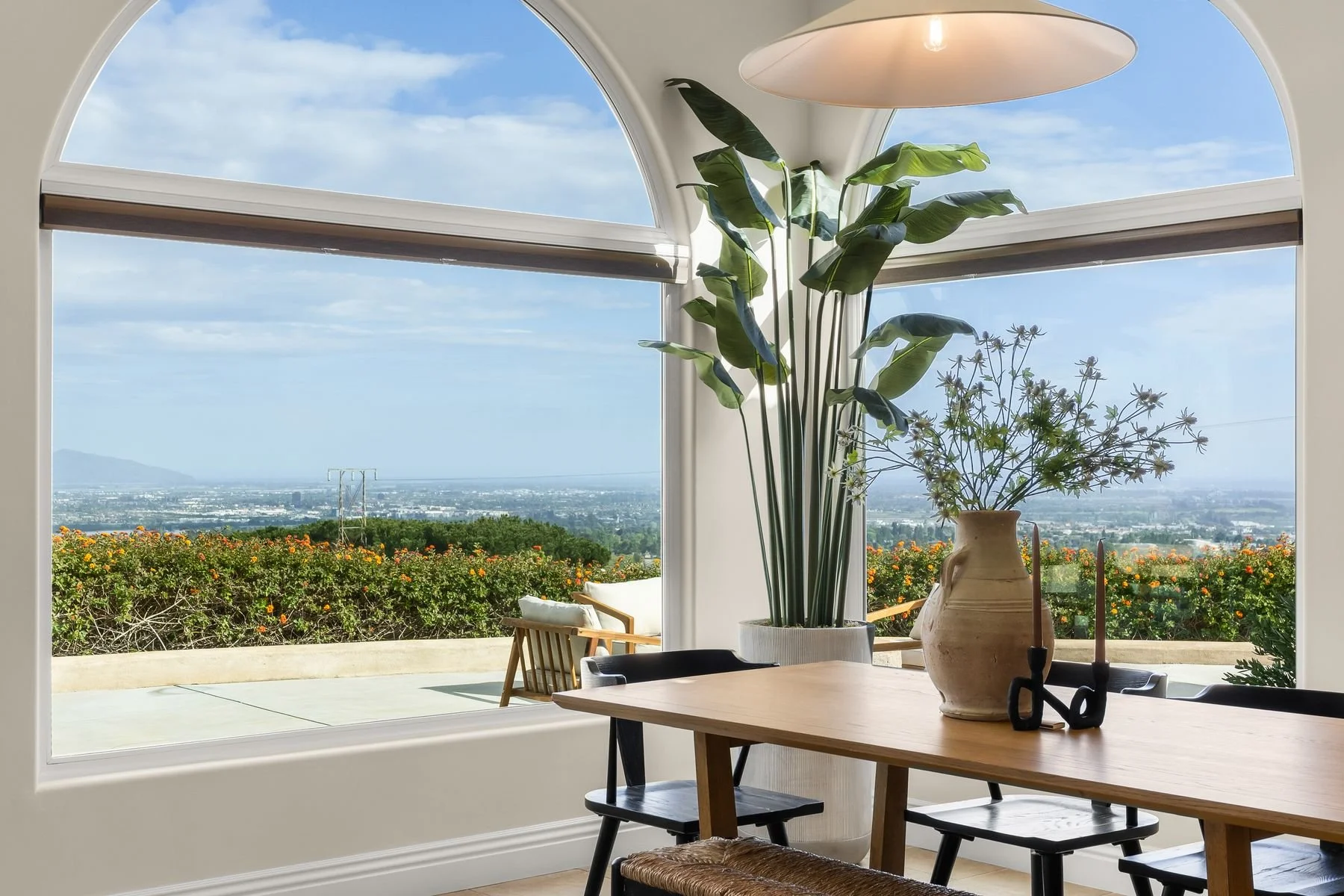 Dining room with large arched window overlooking a garden with orange flowers, bushes, and a distant view of mountains and cityscape under a partly cloudy sky. Interior features a wooden dining table, black chairs, tall green plants, and a beige vase