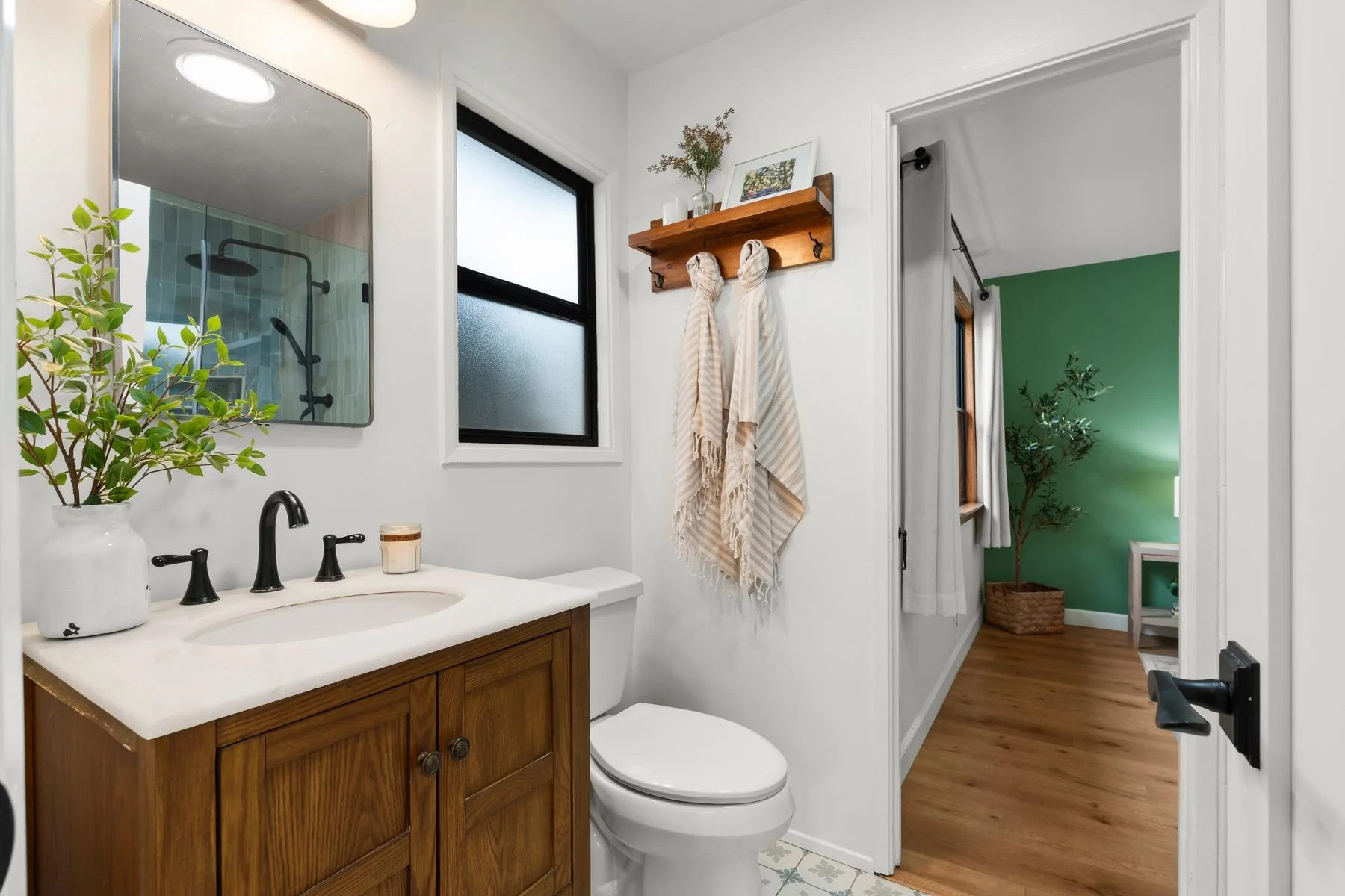 A small bathroom with a wooden vanity, a white countertop, a potted plant, a bathroom mirror, a black faucet, towels hanging on a wooden shelf, a window, a toilet, and a doorway leading to a room with a green wall and a plant.