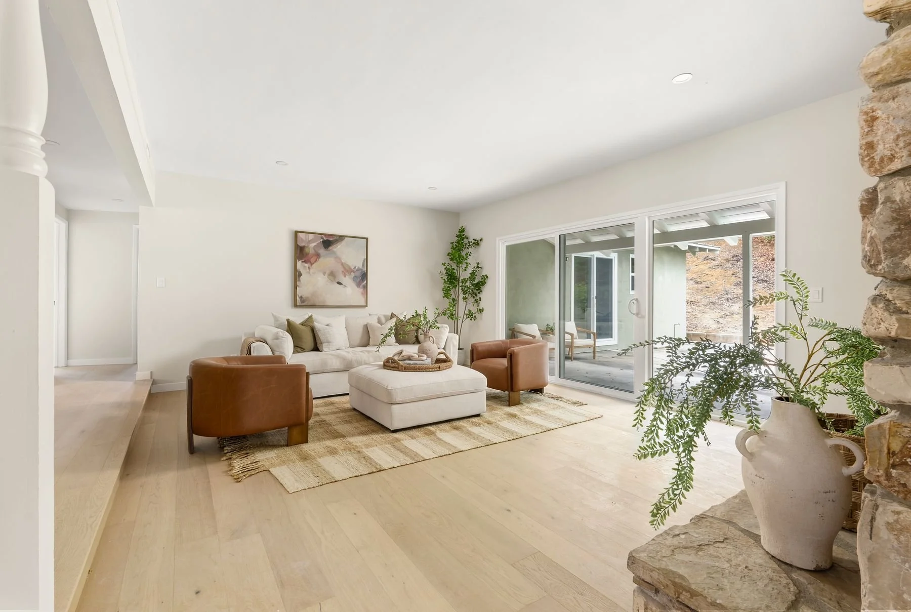 Bright living room with cream sofa, leather armchairs, abstract artwork, sliding glass doors to patio, potted plants, and wooden flooring.