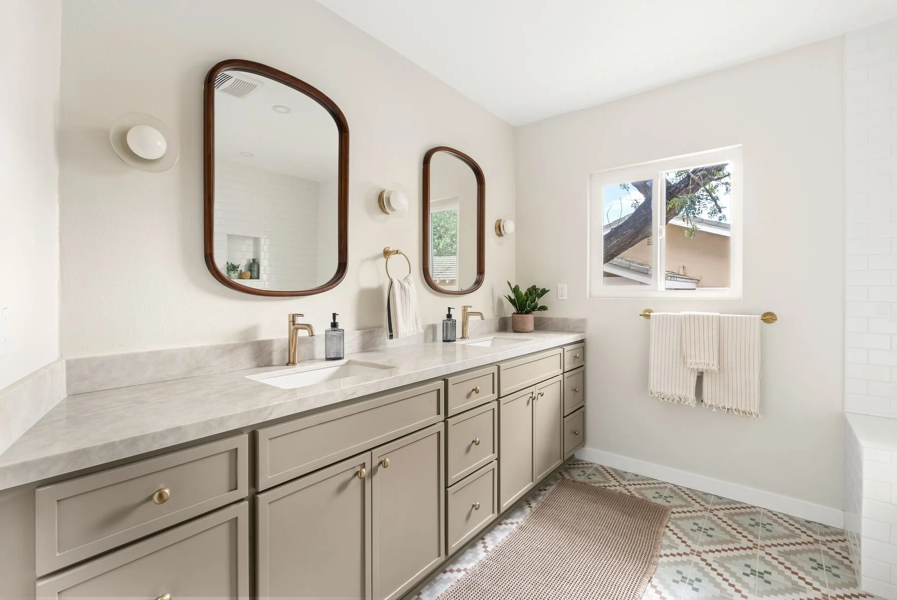 A modern bathroom with a long beige vanity with two sinks, gold faucets, and matching drawer knobs. Above the vanity are two wooden-framed mirrors, each accompanied by a wall-mounted light fixture. There is a window with a view of tree branches and a