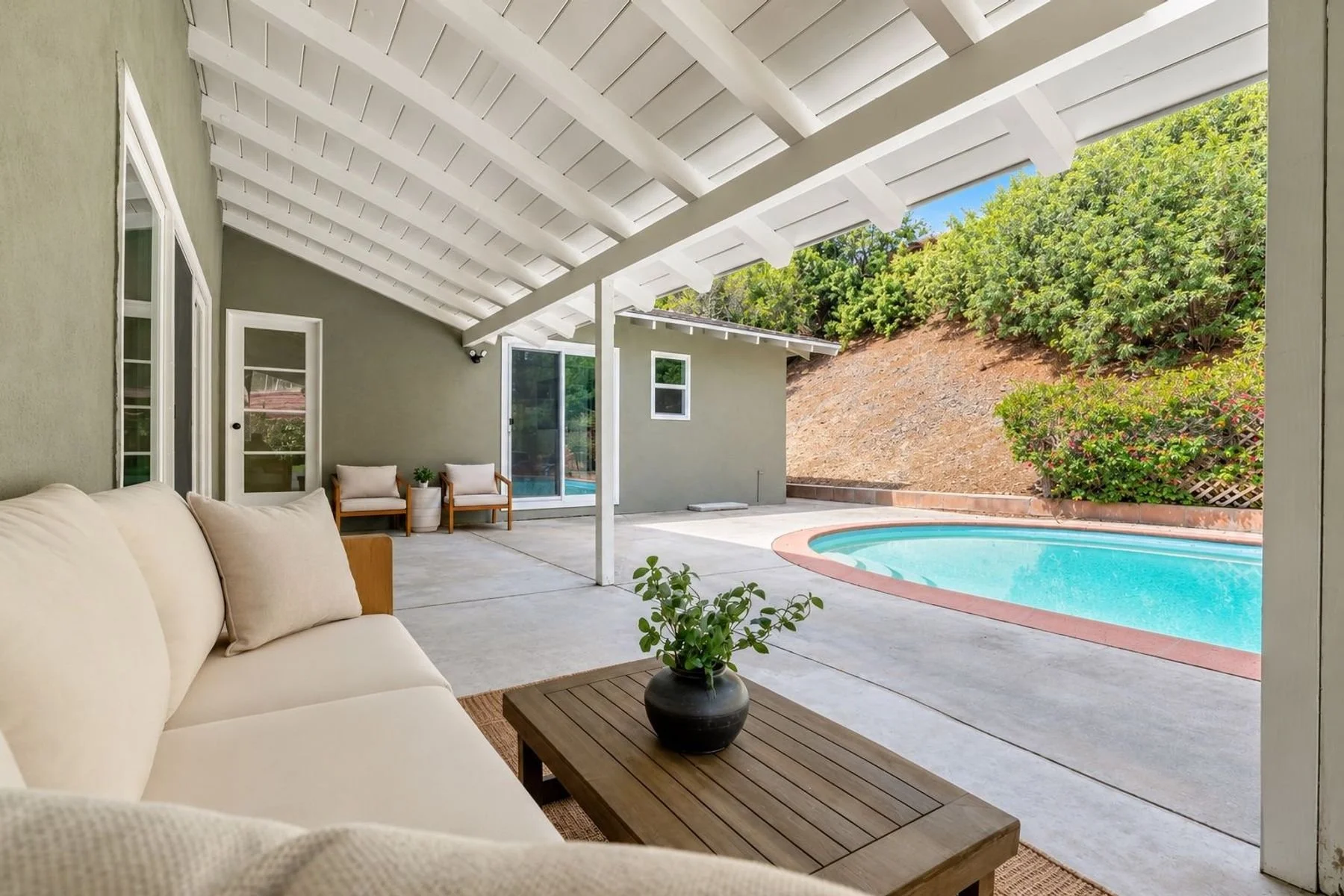 Covered patio area with beige sofa, wooden coffee table with a potted plant, two armchairs with cushions, a swimming pool, and a hillside with trees and bushes outside.