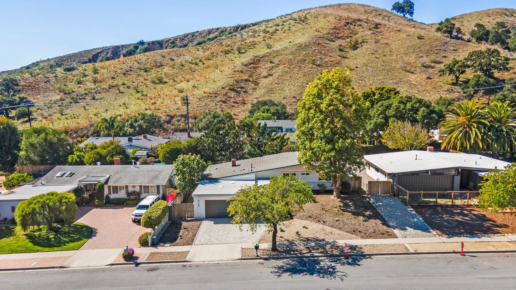 Aerial view of a suburban neighborhood with single-story houses, trees, and a hillside in the background under a clear blue sky.