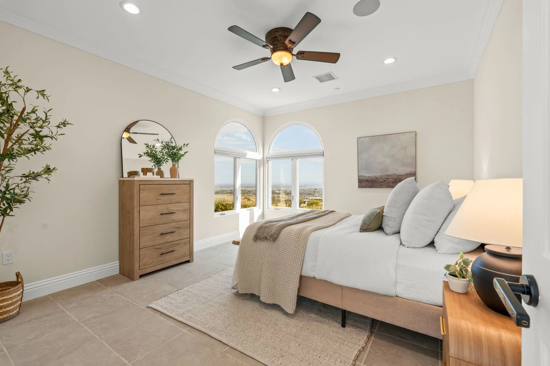 Bright bedroom with large arched windows showing a city view, white bed with pillows and beige blanket, wooden dresser with mirror, potted plants, and a ceiling fan.
