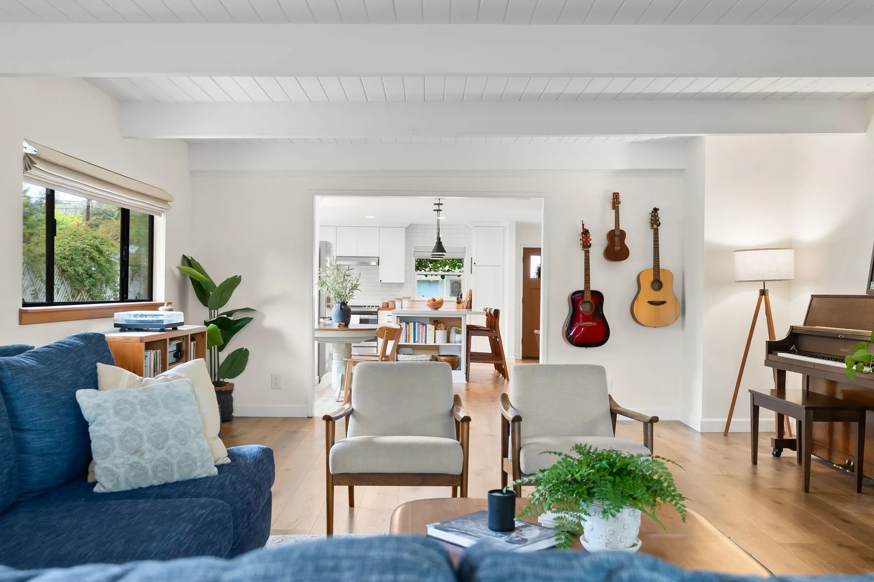 A cozy living room with a blue sofa, white pillows, and a wooden coffee table with a potted fern. There are large windows, a plant, and three beige chairs facing the window. The wall has three guitars hanging, a piano on a stand with a lamp, and a di