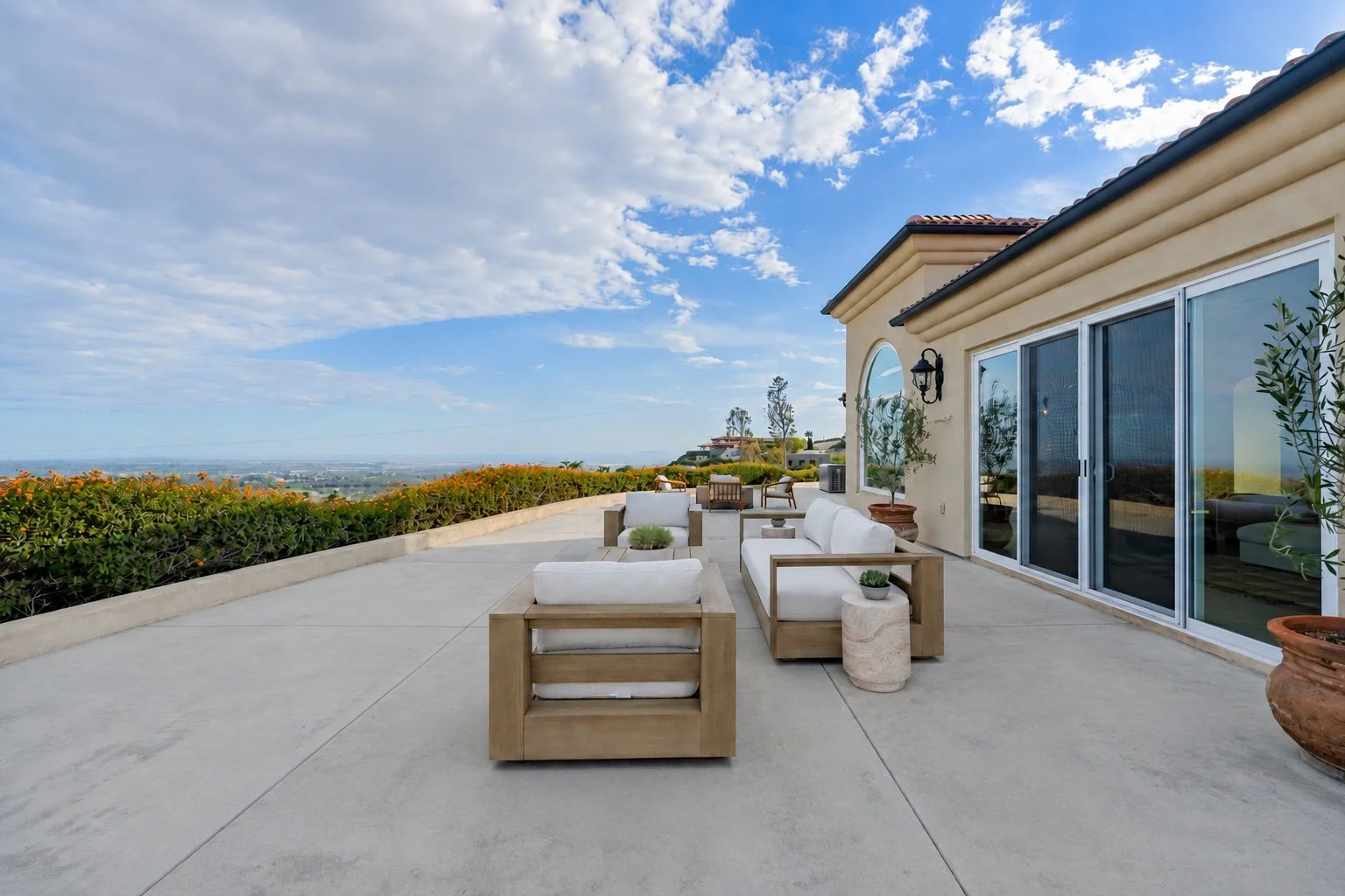 An outdoor patio with modern furniture, potted plants, and a view of a scenic landscape and blue sky with clouds.