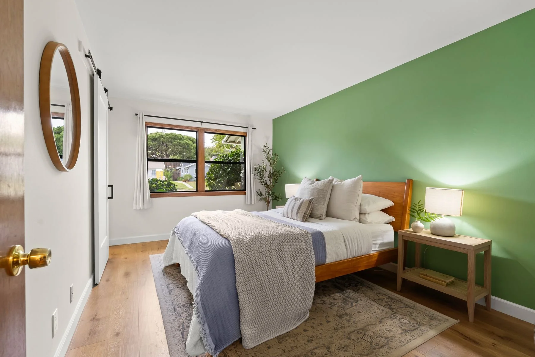 A cozy bedroom with a green accent wall behind a wooden bed frame. The bed has white and beige linens, pillows, and a textured throw blanket. A wooden nightstand with a lamp, small plant, and vase is beside the bed. There is a large window with white