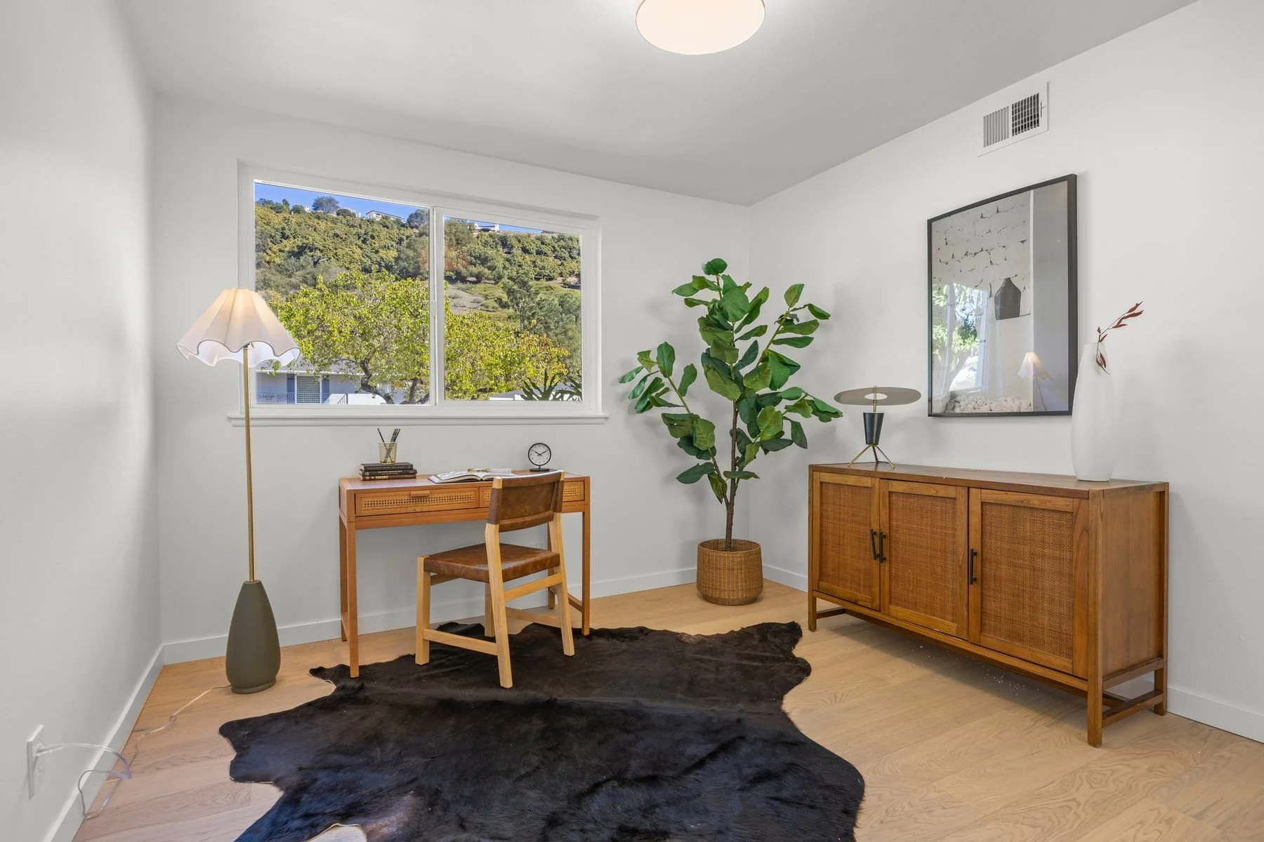 A cozy home office corner with a wooden desk and chair, a tall floor lamp, a large potted ficus, and a wooden cabinet. The room has white walls, a window showing a green hillside, and a black animal hide rug on the light wood floor.