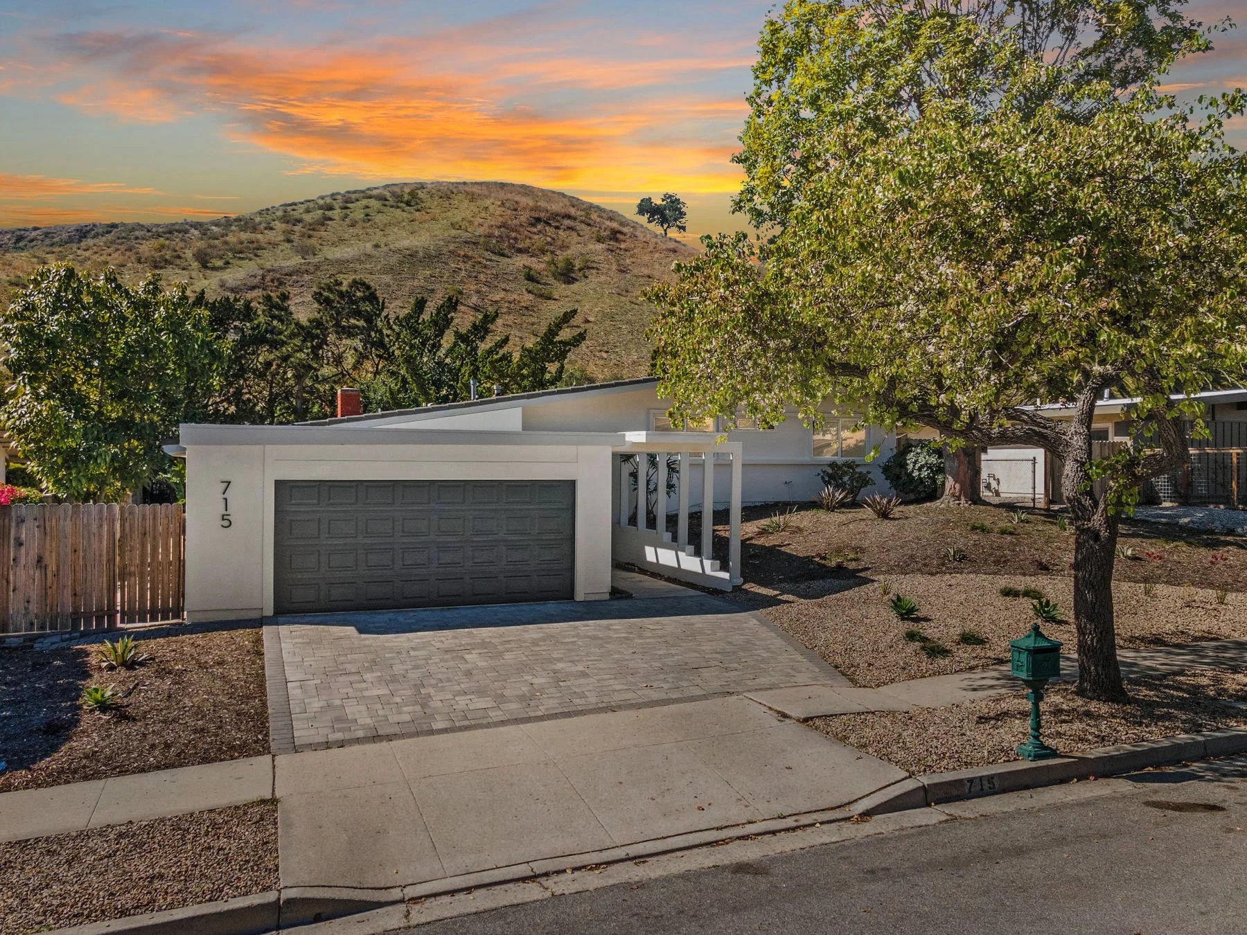A modern suburban house with a gray garage door, a small driveway paved with bricks, a sidewalk, a tree with green leaves, and a mailbox. In the background, a hill with dry grass and a few trees under a colorful sunset sky.