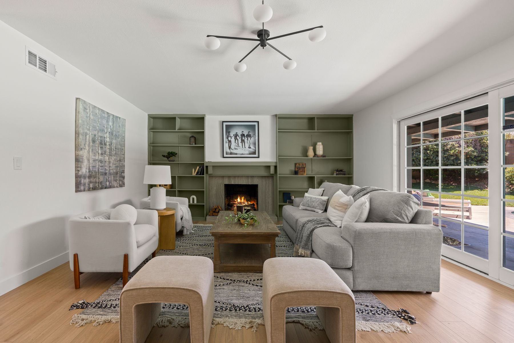 A living room with a gray sofa and white armchair arranged around a wooden coffee table. There is a fireplace with a fire, and built-in shelves on either side with decorative items. Large sliding glass doors lead outside to a patio and garden. A blac