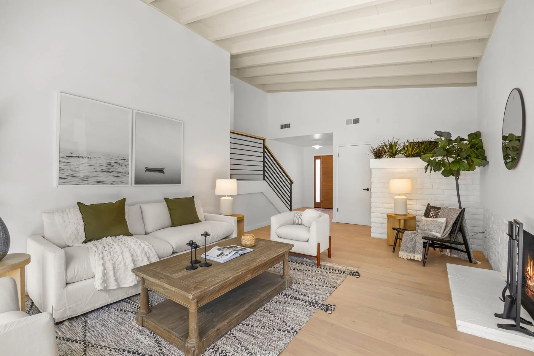 Modern living room with white sofa, wooden coffee table, armchair, and a fireplace with a large plant on top. Black and white artwork hangs on the wall, and there are lamps providing warm lighting.