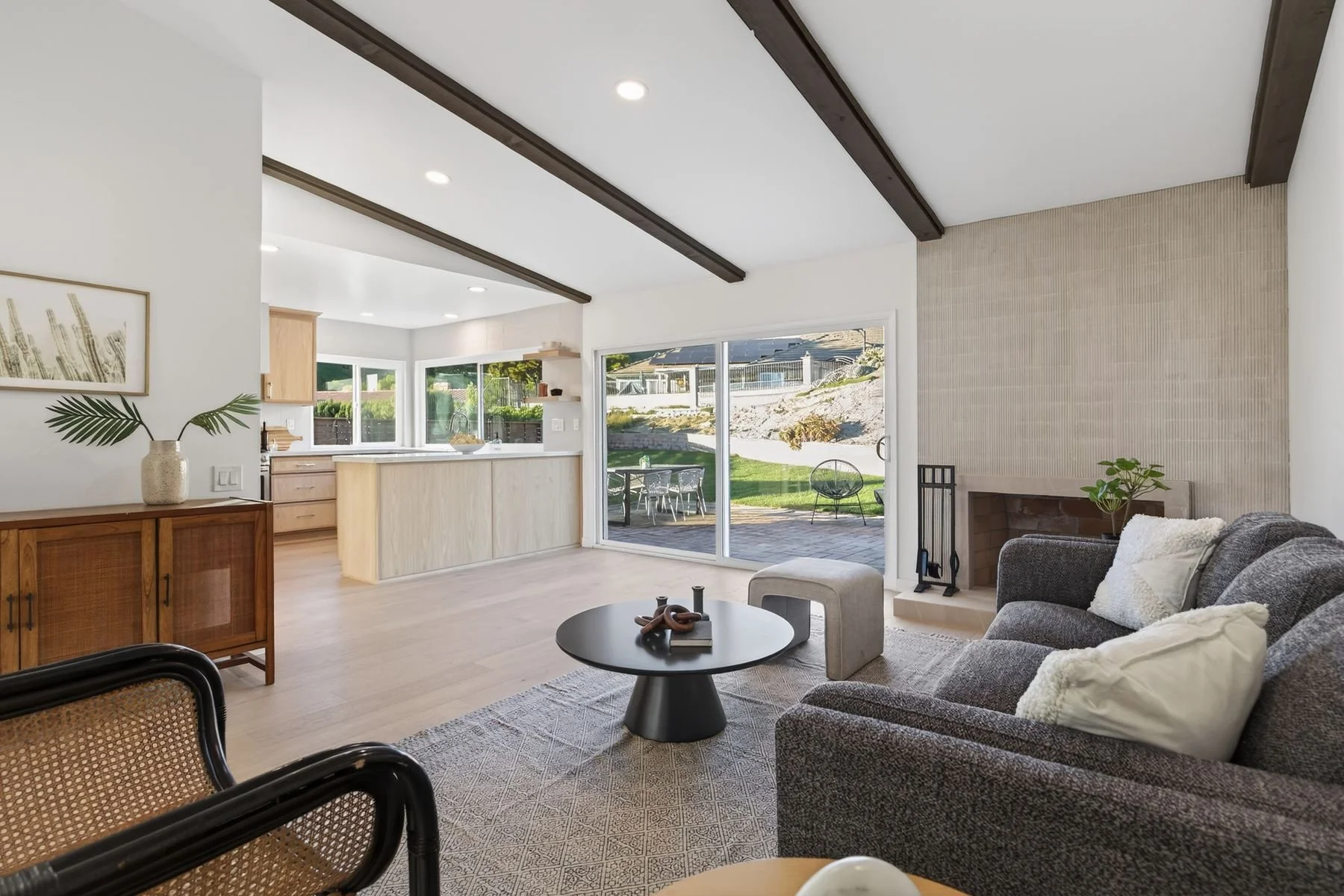 Living room with gray sofa and pillows, round black coffee table, large window with sliding glass door leading to outdoor patio, kitchen with light wood cabinets and white countertops, fireplace with beige textured wall, ceiling with dark wooden beam