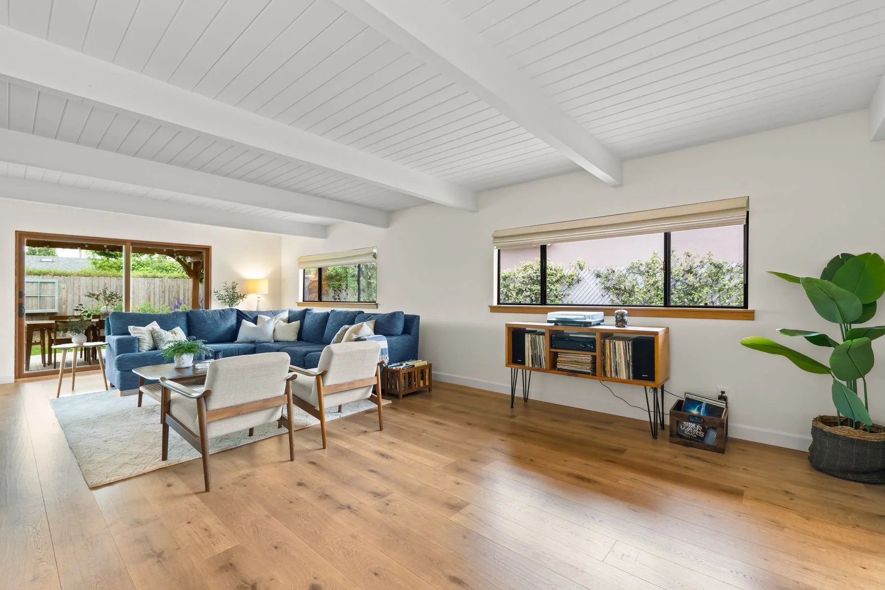Living room with white wooden ceilings, large windows, and a sliding glass door leading outside. It has a blue sectional sofa with throw pillows, two light-colored armchairs, a wooden coffee table with a plant, a wooden media console with records and