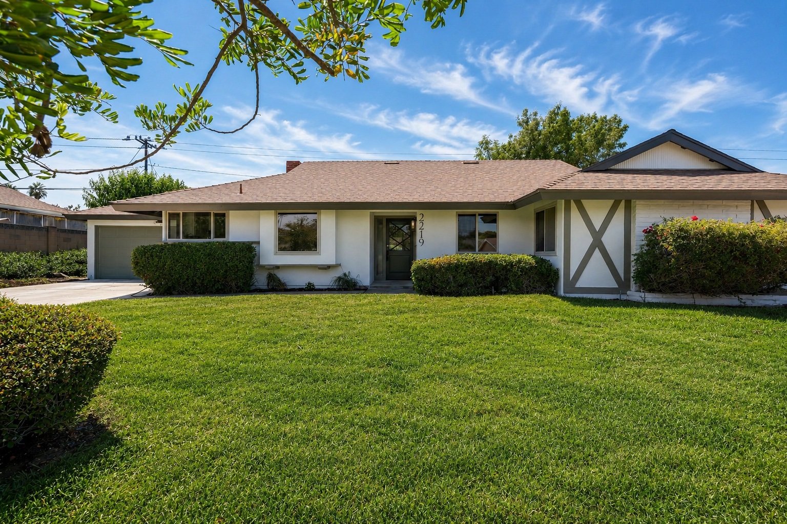 A single-story white house with a brown roof, large front windows, and a front door with house number 2219. The house has a well-maintained green lawn, shrubs, and trees, with a bright blue sky and scattered clouds overhead.