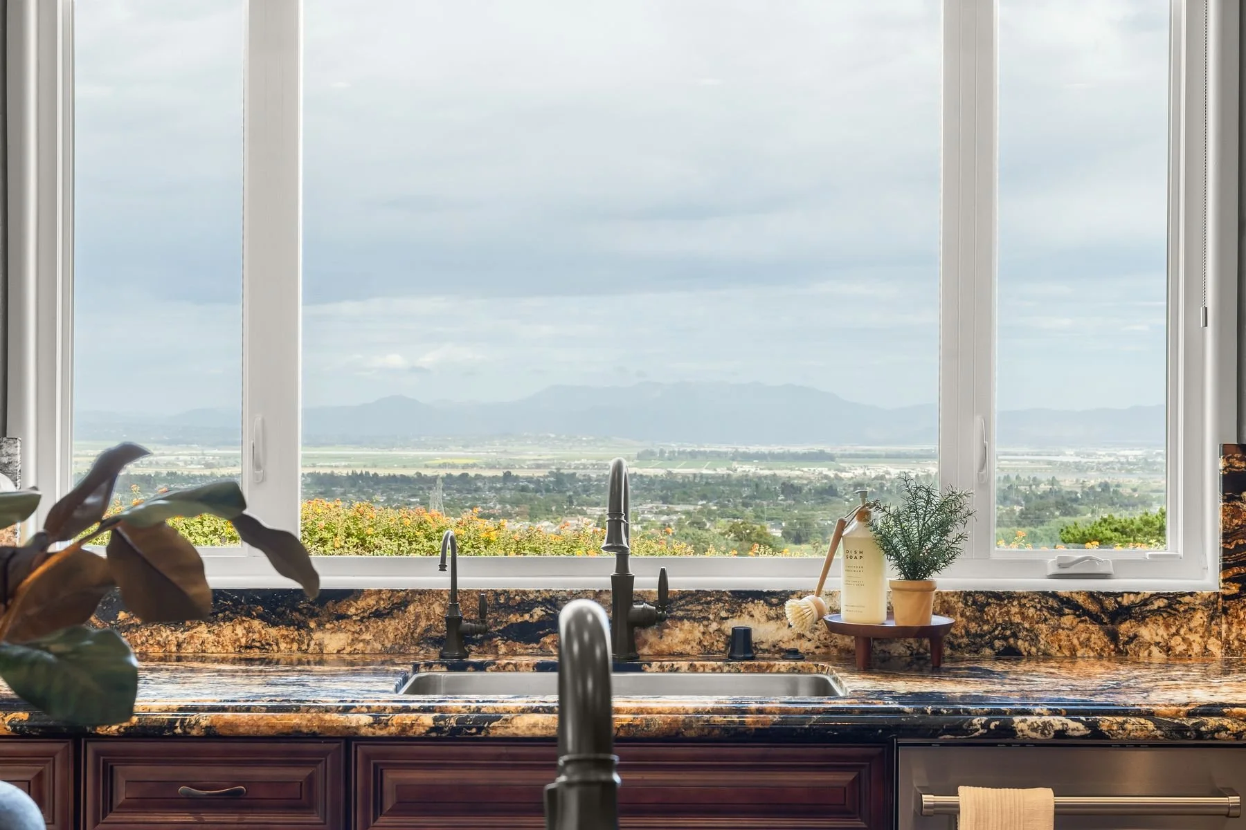 Kitchen sink area with a large window overlooking a scenic landscape of mountains, fields, and sky. The countertop is granite with a faucet, soap dispenser, and decorative plants, creating a cozy and inviting atmosphere.