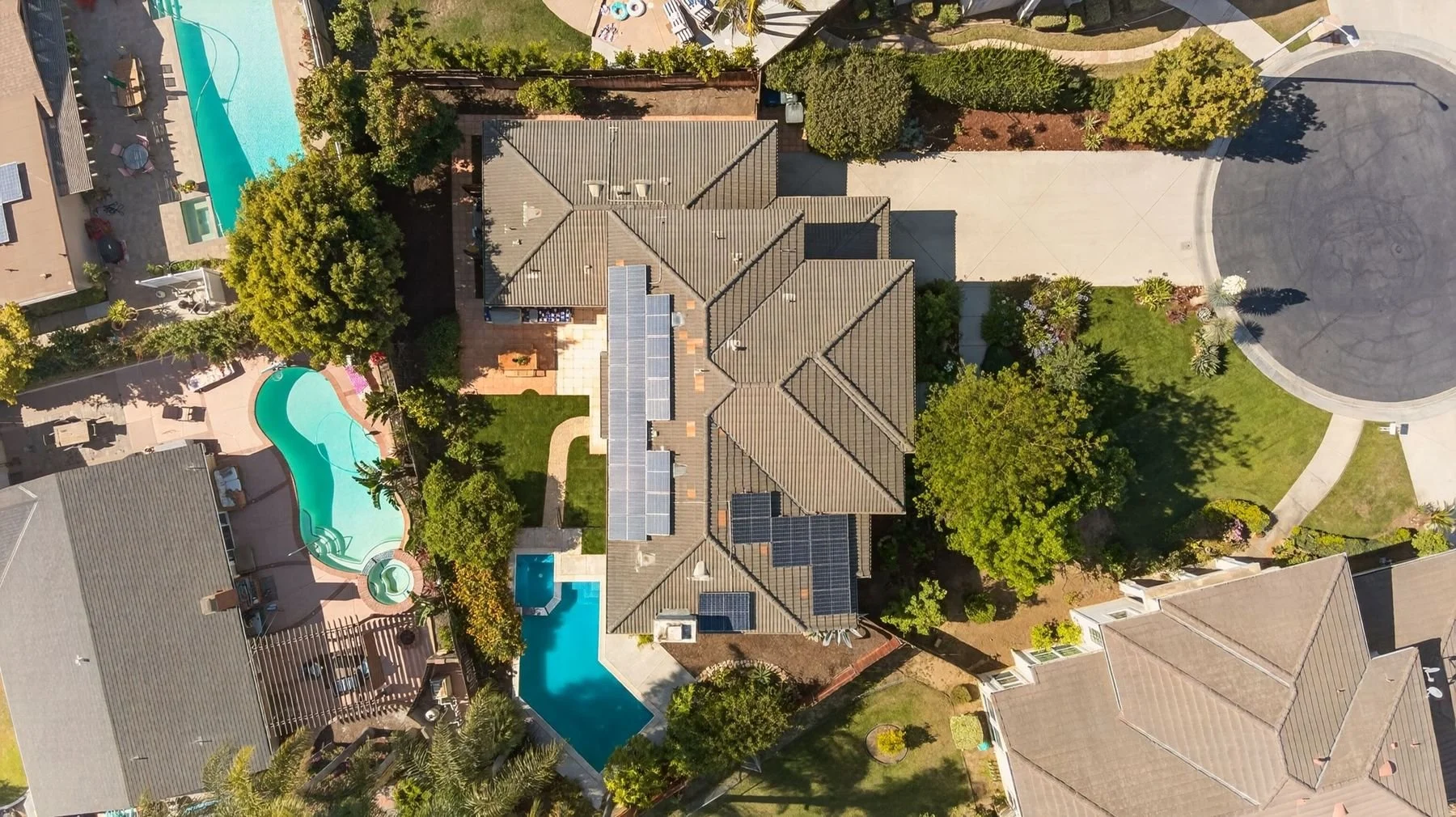 Aerial view of a residential neighborhood showing houses, garages, and yards with swimming pools, trees, sidewalks, and a street.