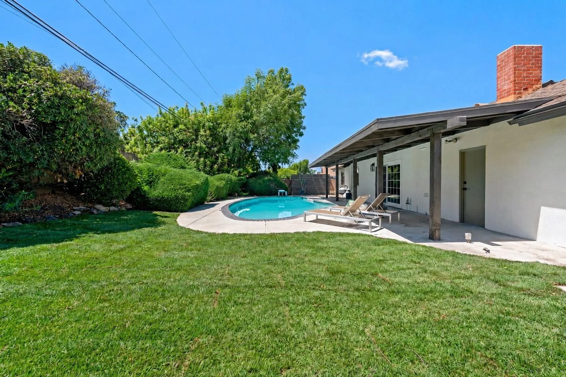 A backyard with a grassy lawn, a small round swimming pool, and four loungers next to a house with a covered patio. There are trees, bushes, and a clear blue sky with a few clouds.