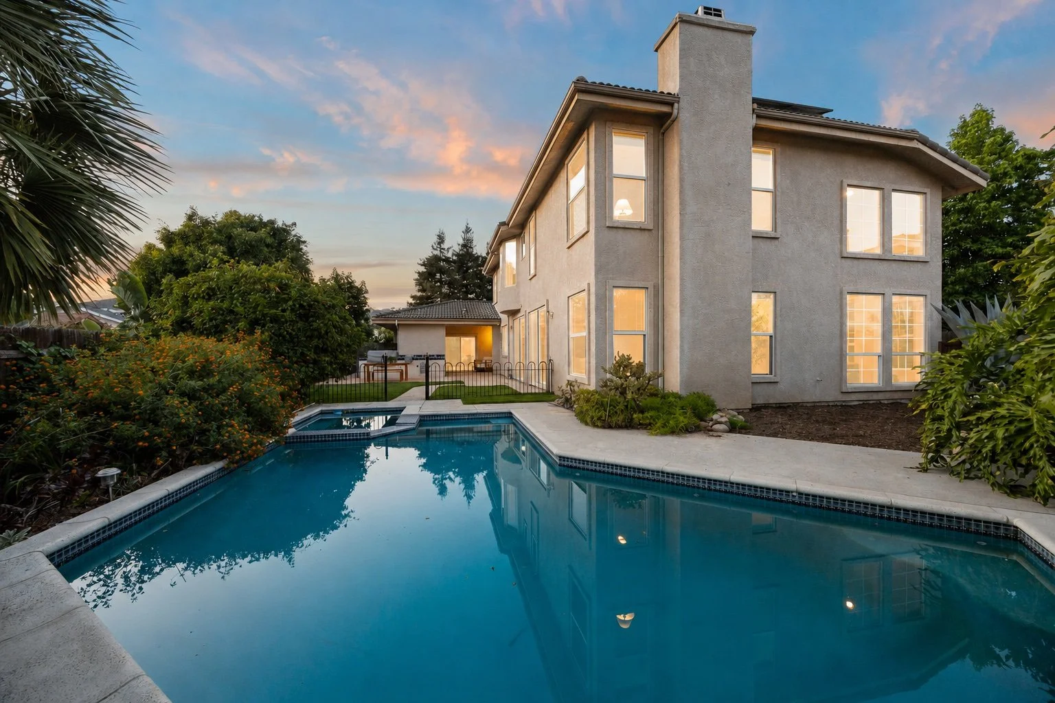 Backyard view of a two-story house with large windows, a swimming pool, and surrounding greenery at sunset.