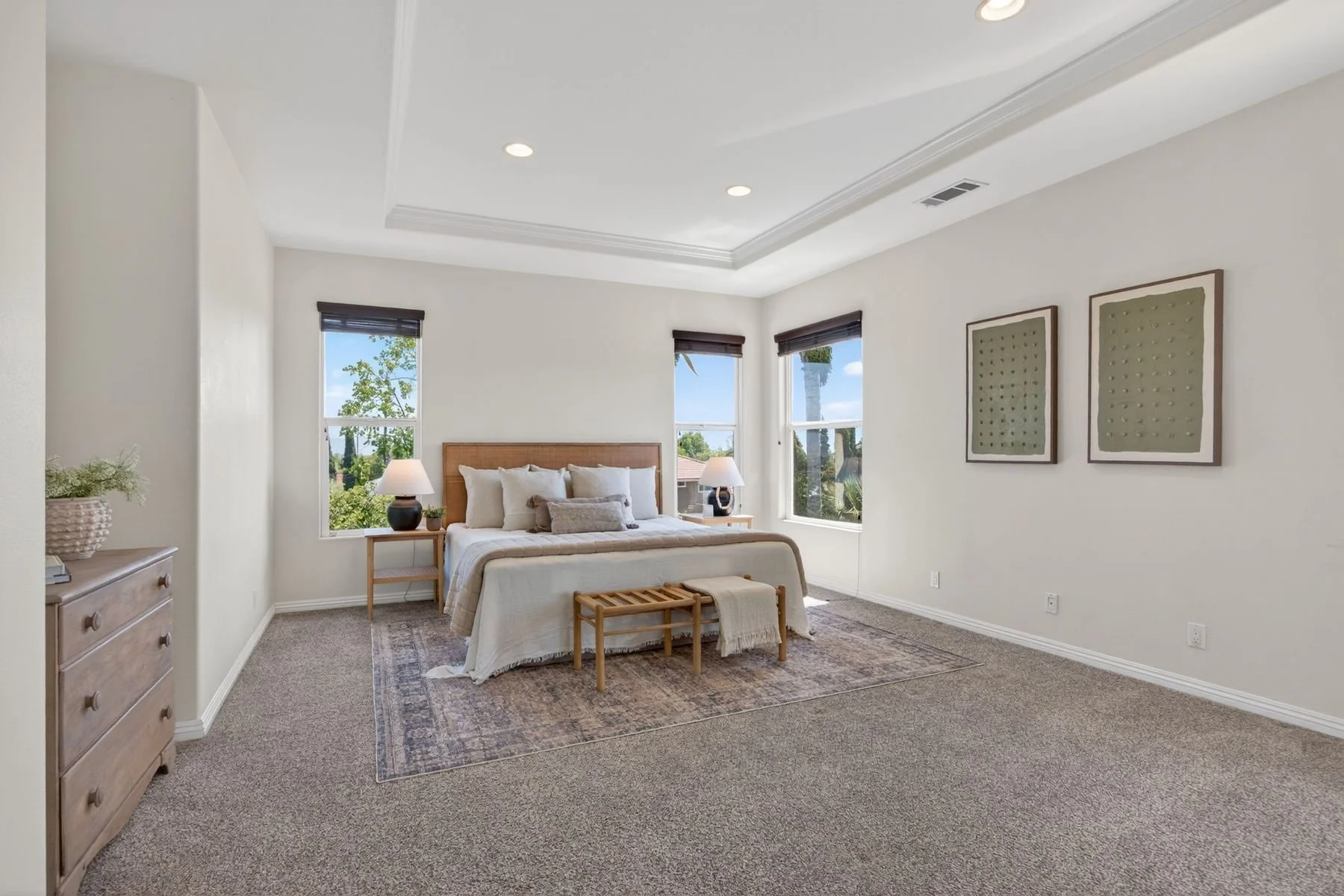 A bedroom with a bed, two nightstands with lamps, and three windows showing trees outside, decorated with two framed abstract artworks on the wall.