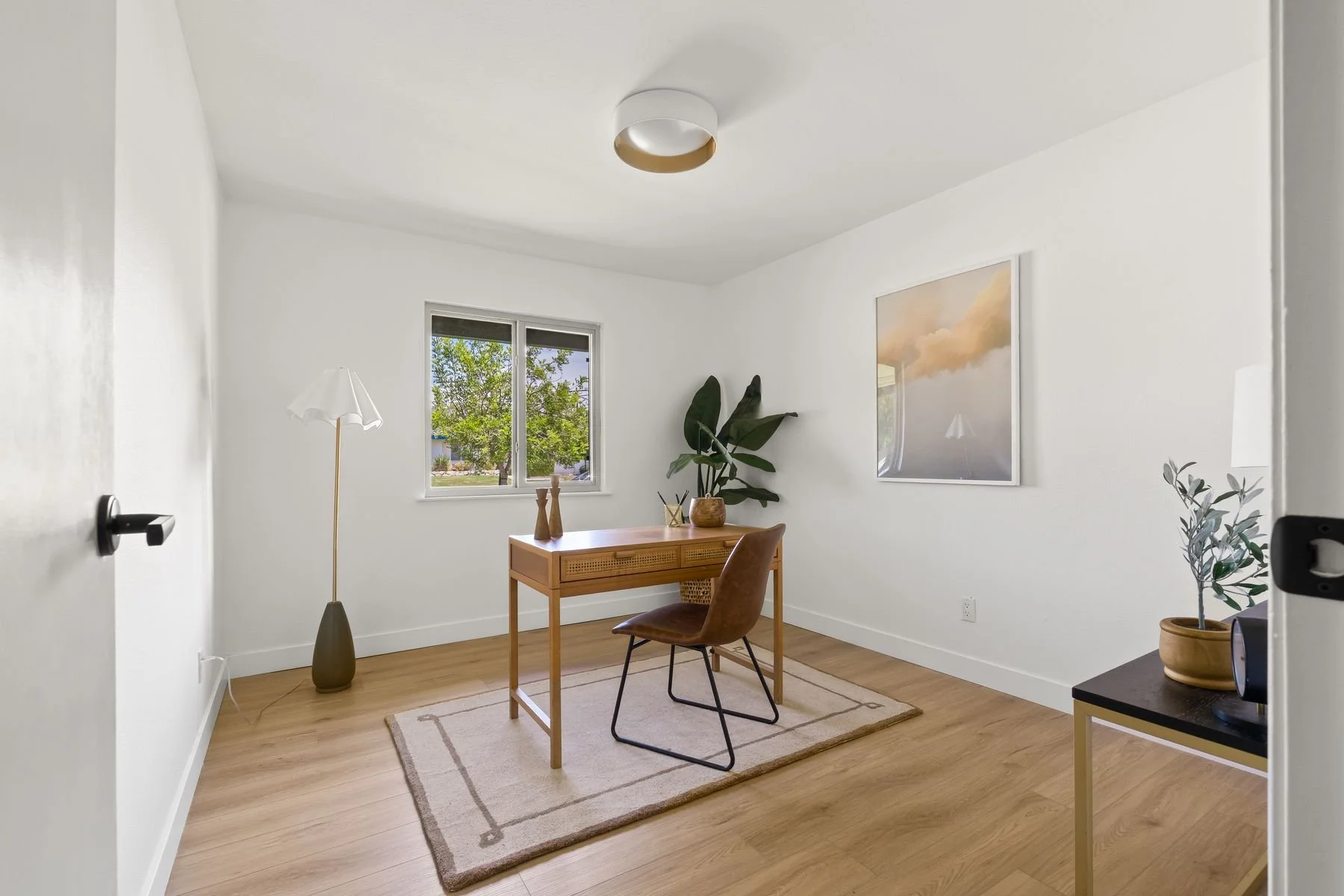 A minimalist home office with white walls, a wooden desk, a leather chair, a window with outside greenery, a standing lamp, potted plants, and a framed art print of clouds.