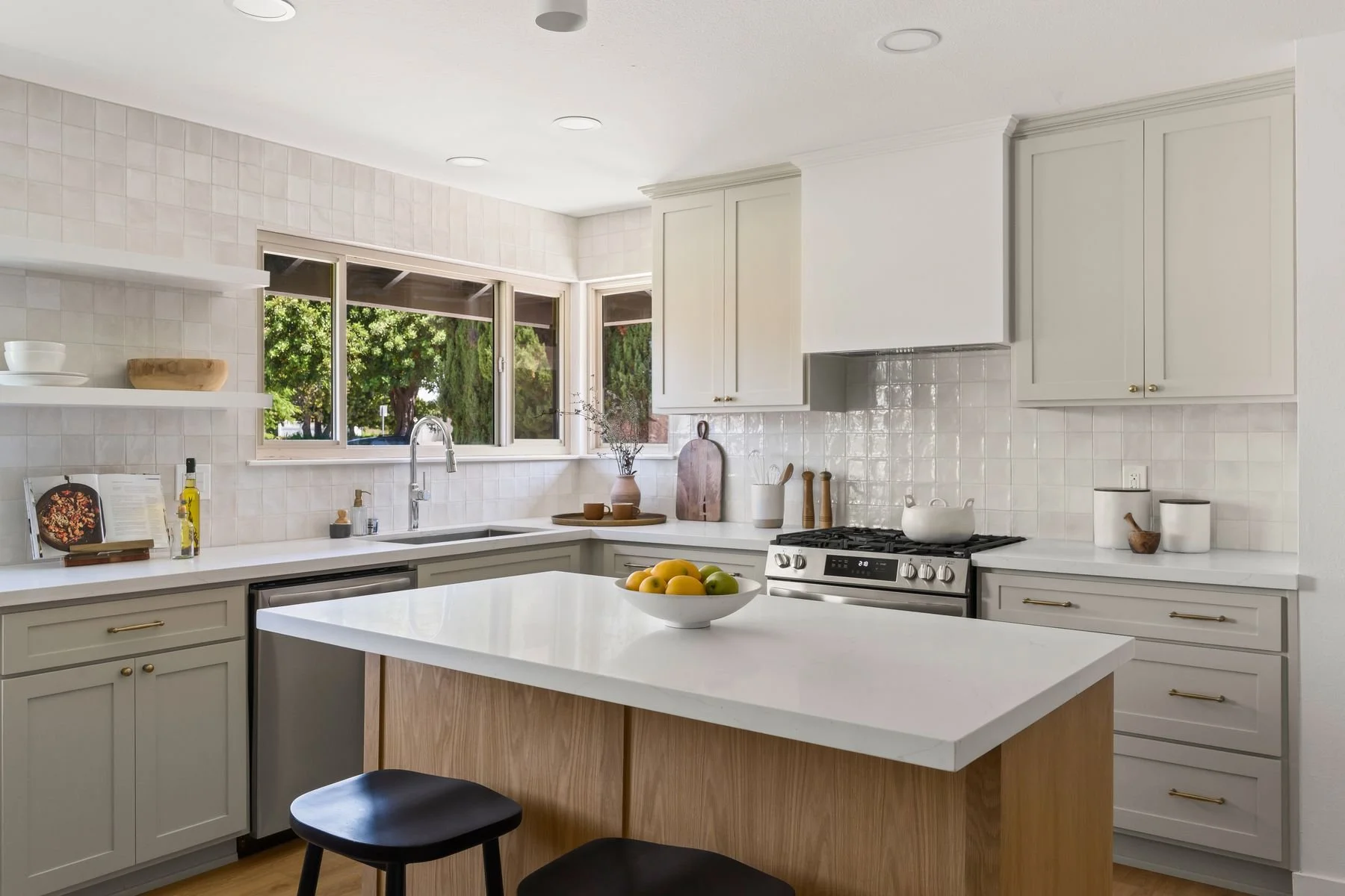 Modern white kitchen with open window, white countertops, and an island with a fruit bowl. Includes a stove, cabinetry, and decorative items like a cutting board, bowls, and a vase with branches.