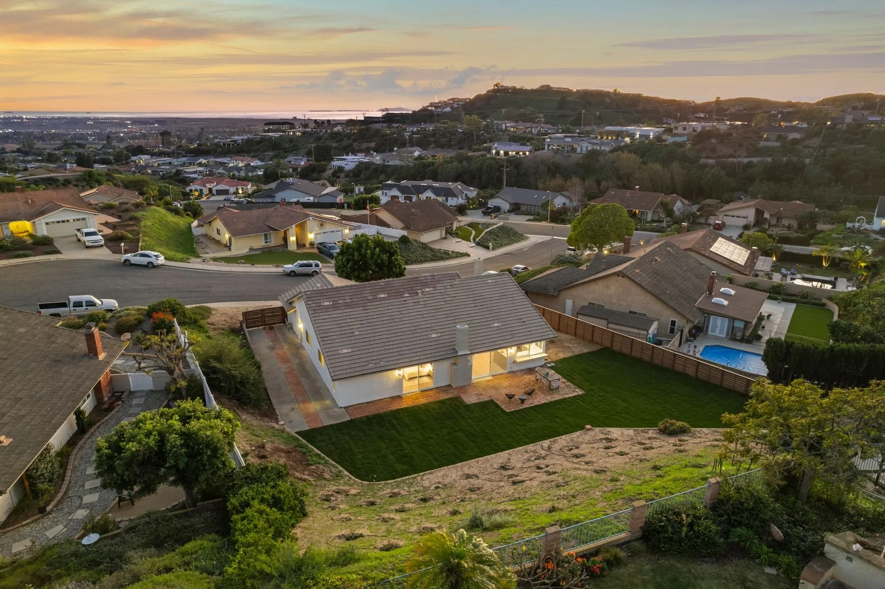 Aerial view of a suburban neighborhood at sunset featuring houses, driveways, a backyard with a swimming pool, well-manicured lawns, and hills in the background.