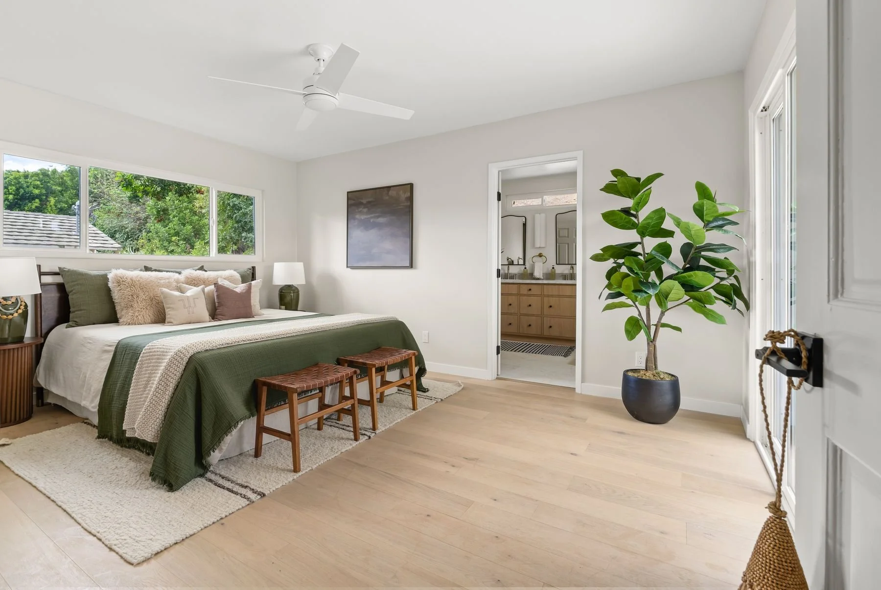 A bedroom with a large bed dressed in white and green bedding, decorated with multiple pillows. On each side of the bed are nightstands with lamps. There is a large window behind the bed showing outdoor greenery. A potted plant is placed near the doo